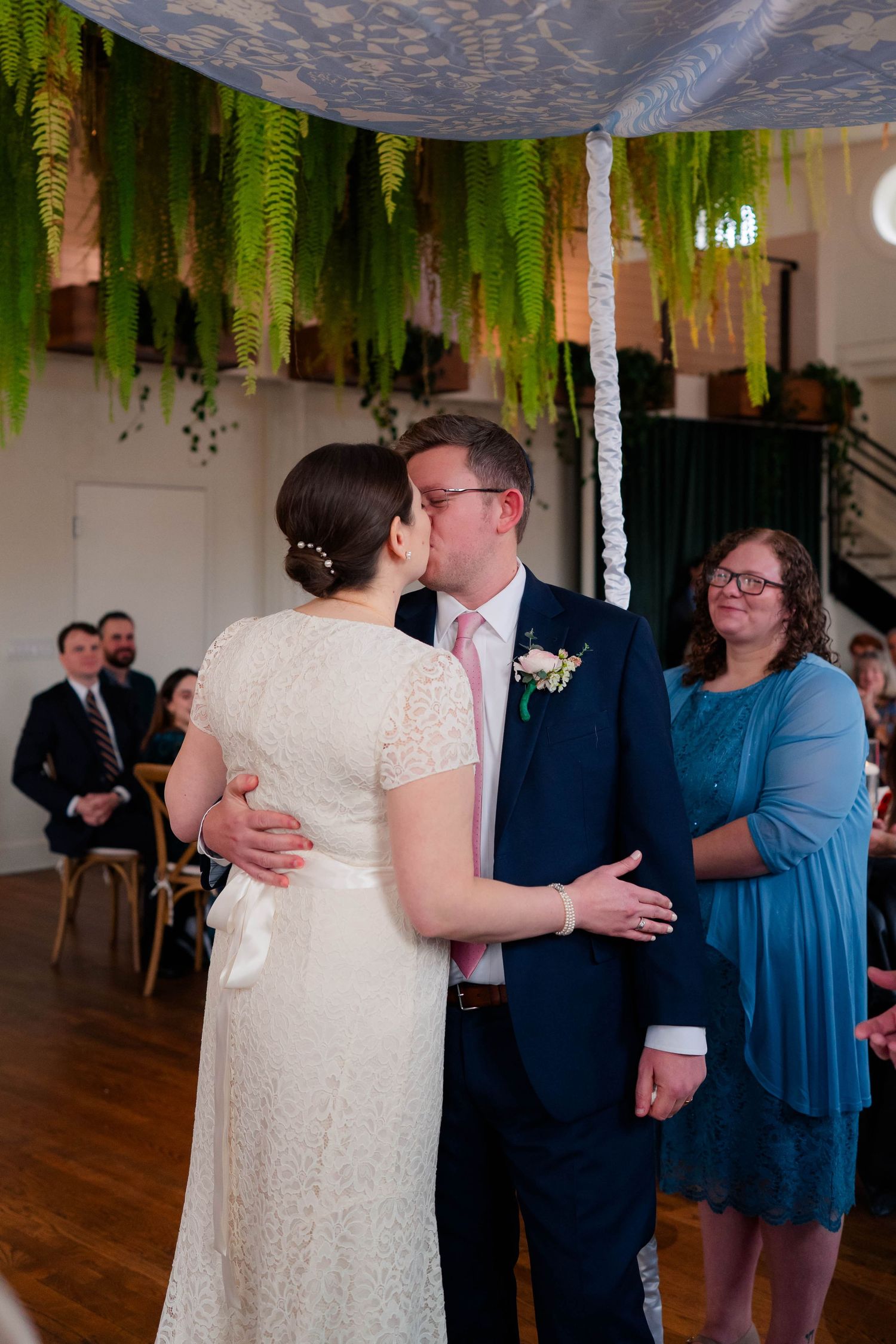 Bride and groom share first kiss under fern-adorned chuppah as wedding party looks on during ceremony.