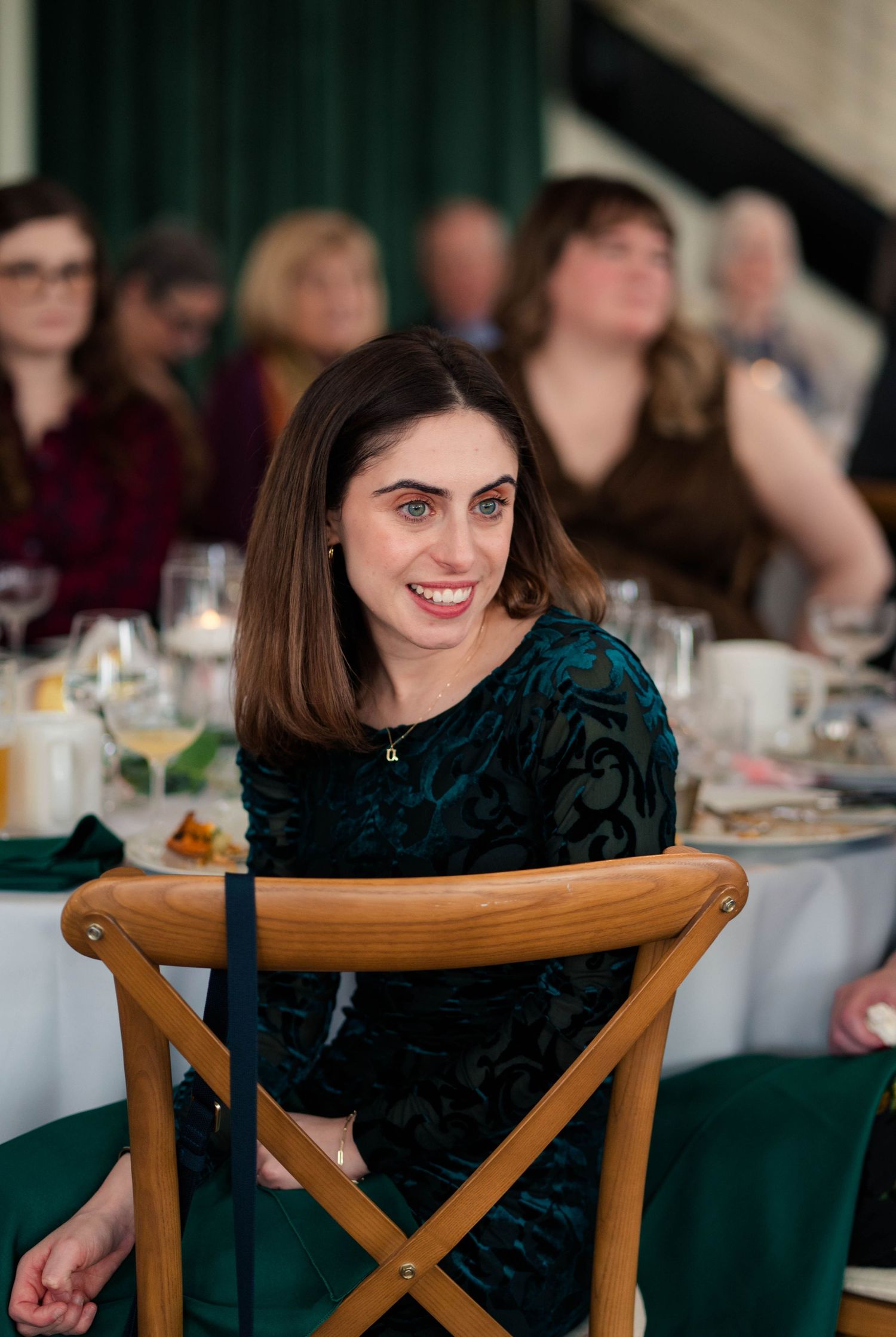 Smiling woman in teal velvet dress seated at wedding reception table with guests in background.