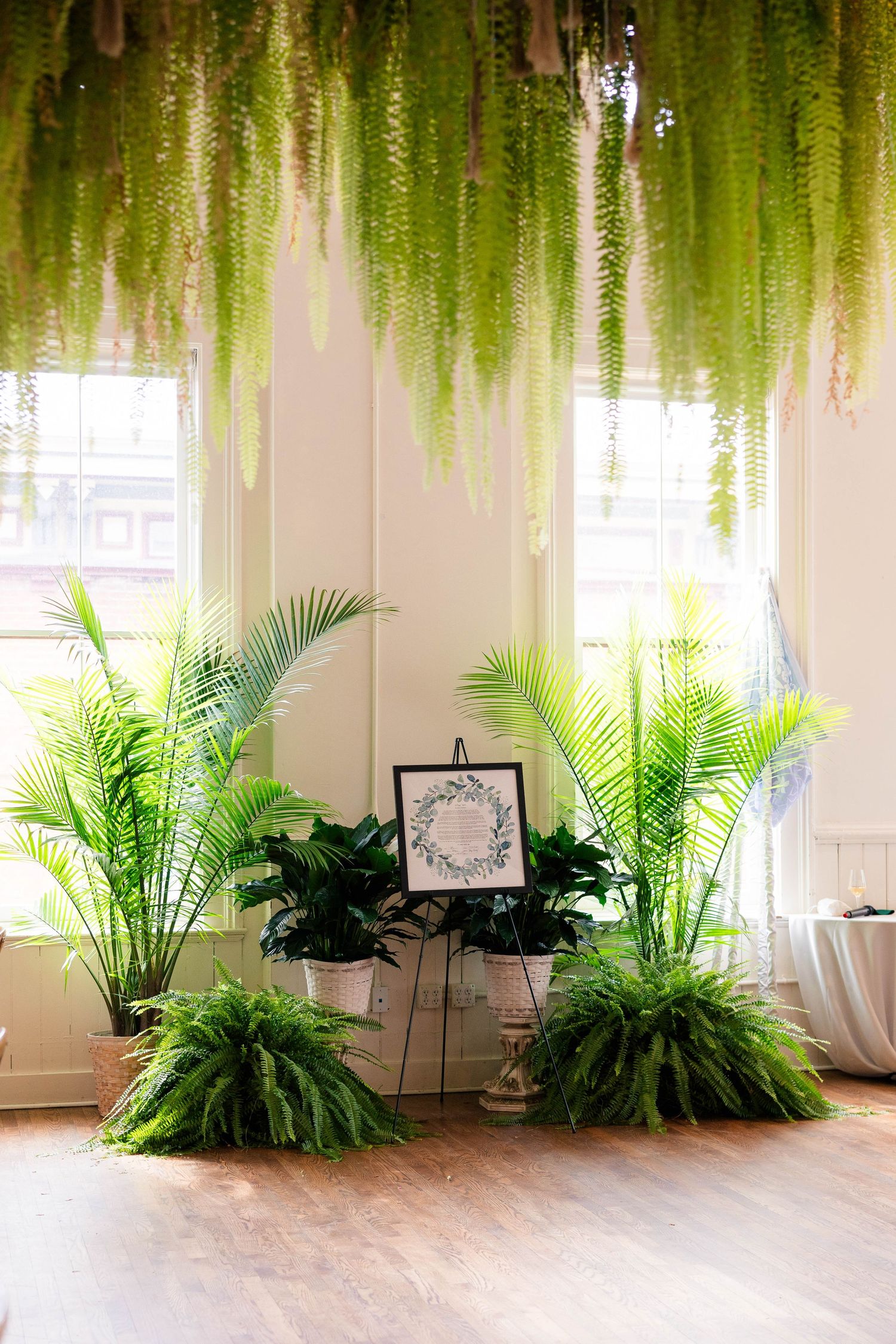 Lush tropical greenery installation with hanging ferns, potted palms, and a framed sign on an easel.