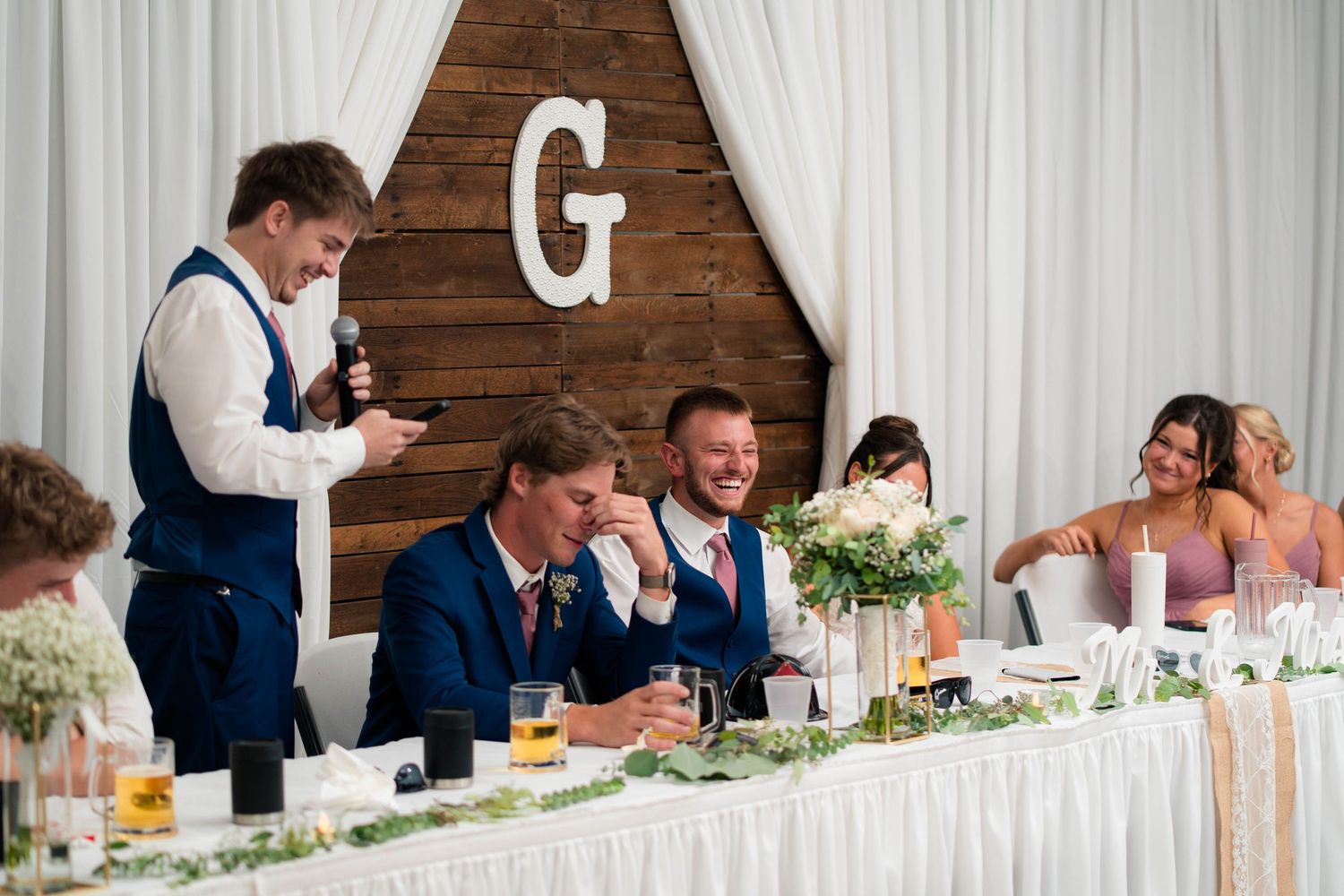 Best man gives toast as groom and bride laugh at elegantly decorated wedding head table with wooden G backdrop.