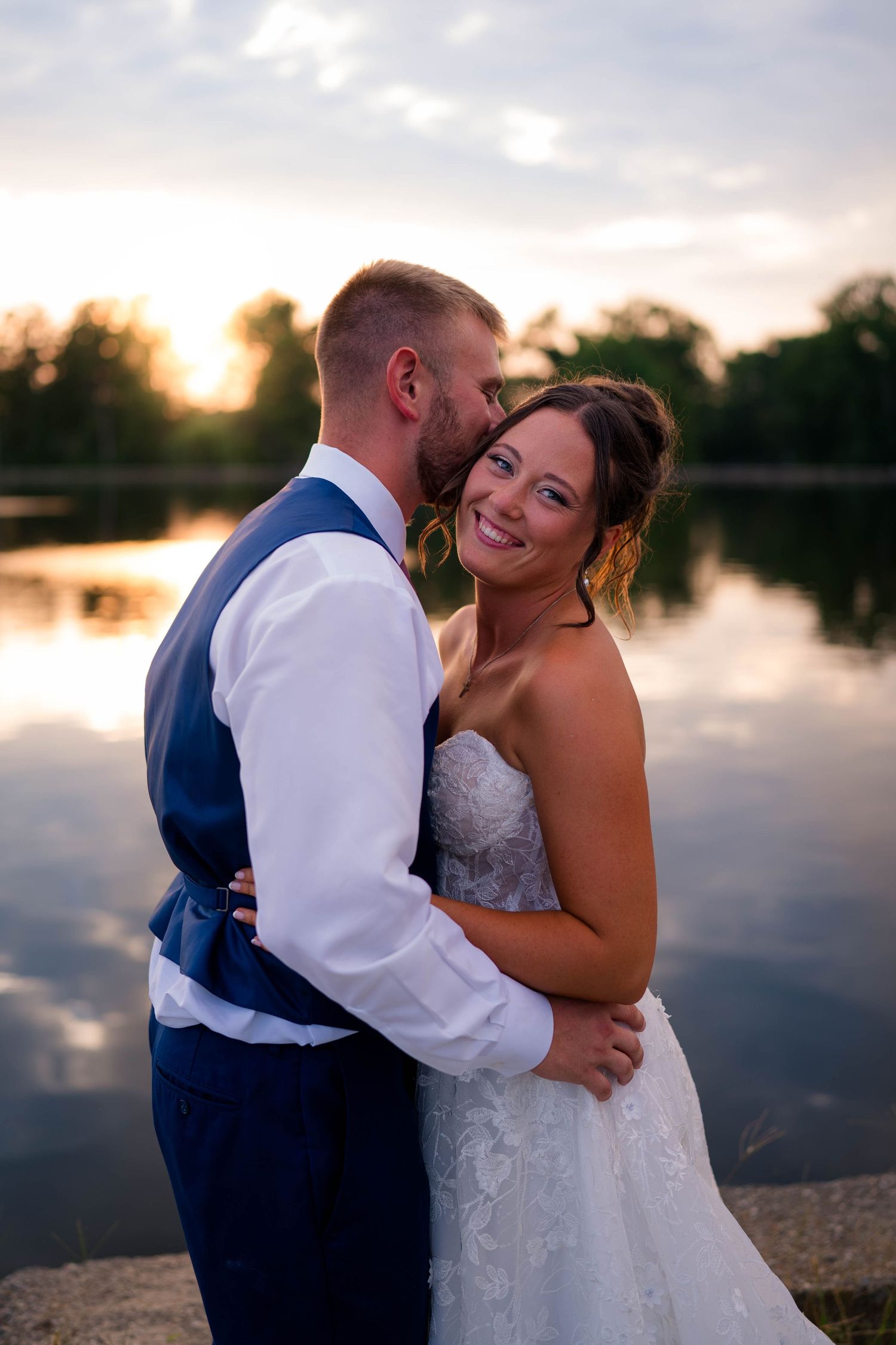 Smiling bride in strapless lace gown leans into groom's embrace by a glowing sunset lake reflection.