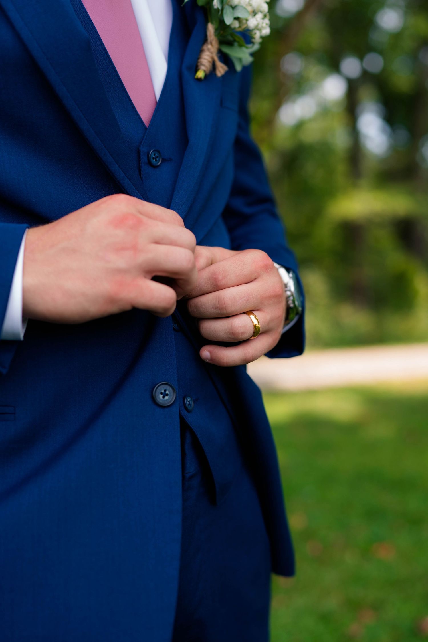 Groom in navy blue three-piece suit adjusting jacket buttons, wearing gold wedding ring and watch with floral boutonniere.