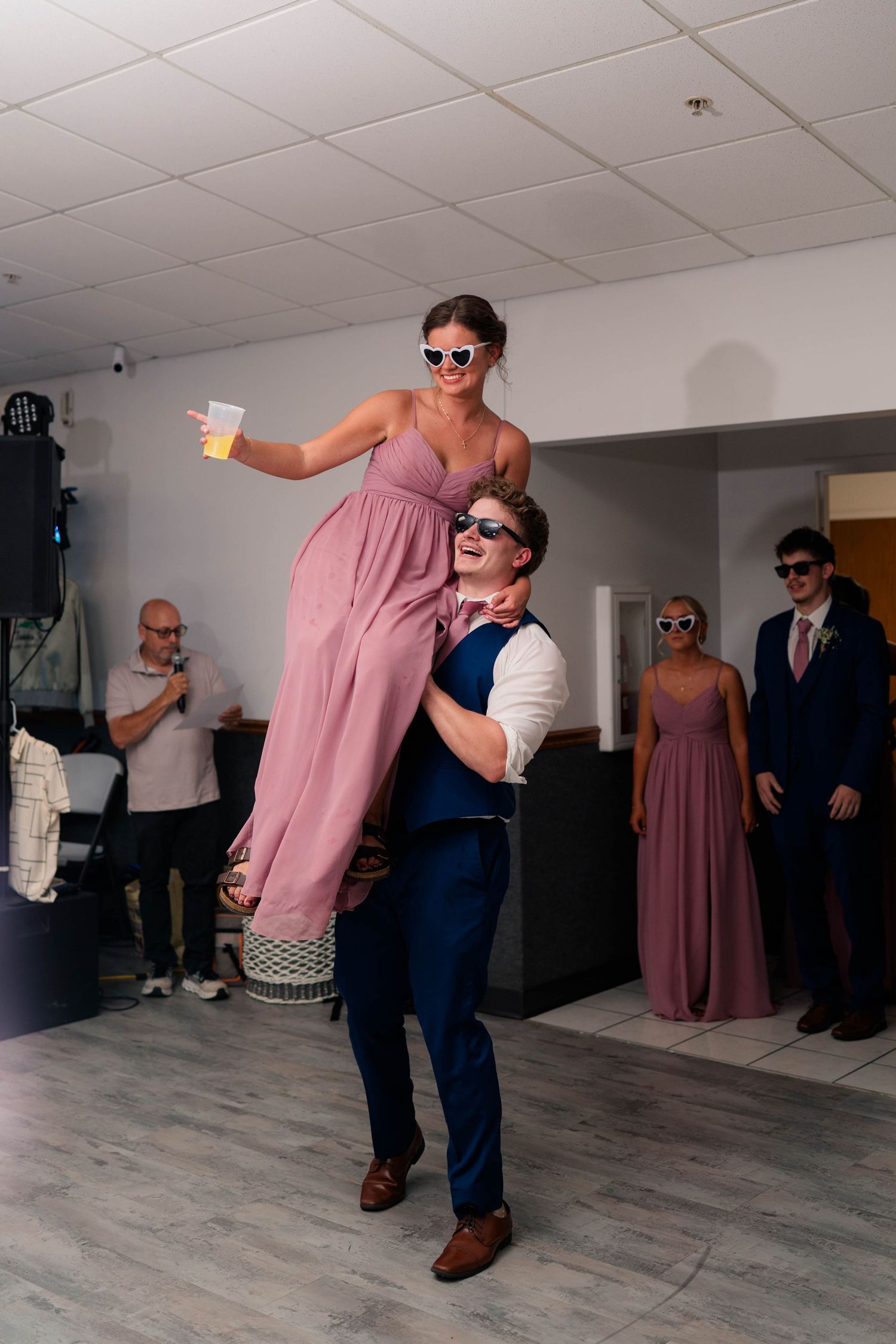 Groomsman in navy suit lifts bridesmaid in pink dress on the wedding reception dance floor.