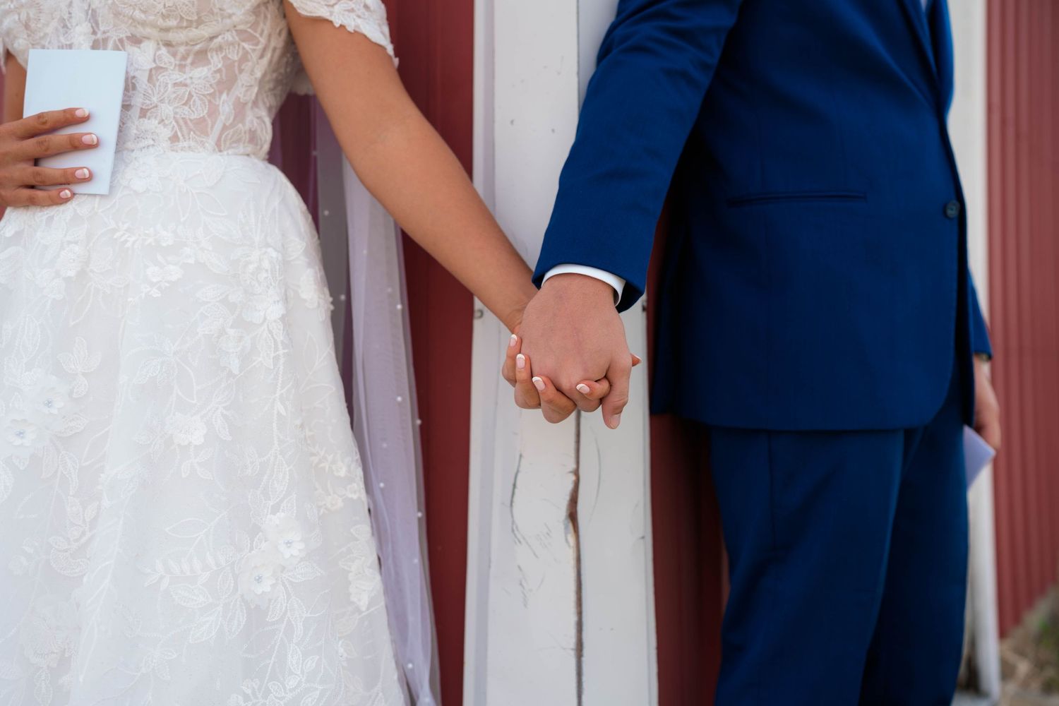 Bride in white lace gown and groom in blue suit holding hands at their wedding ceremony.