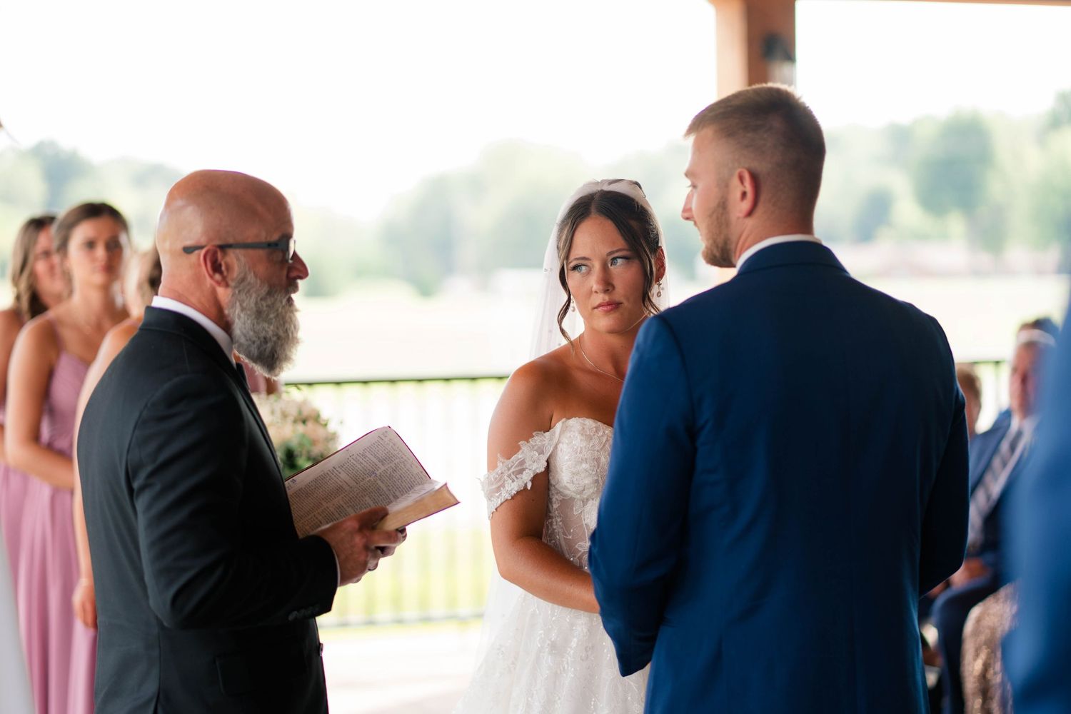 Bride and groom exchange vows with officiant during outdoor wedding ceremony, bride wearing lace gown.