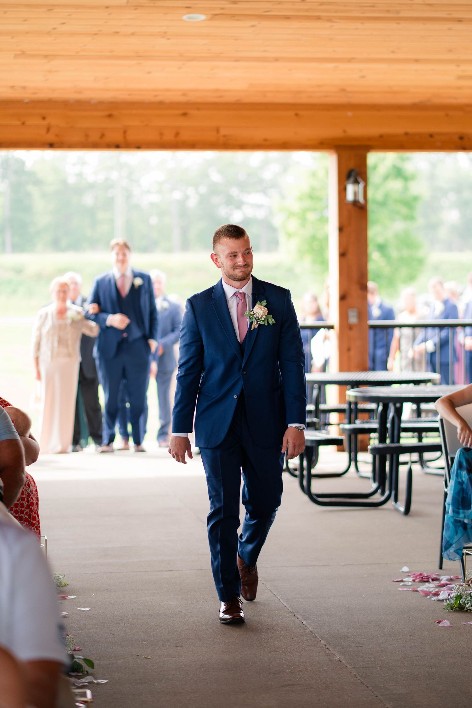 Groom in navy suit walking down aisle at outdoor wedding ceremony pavilion.