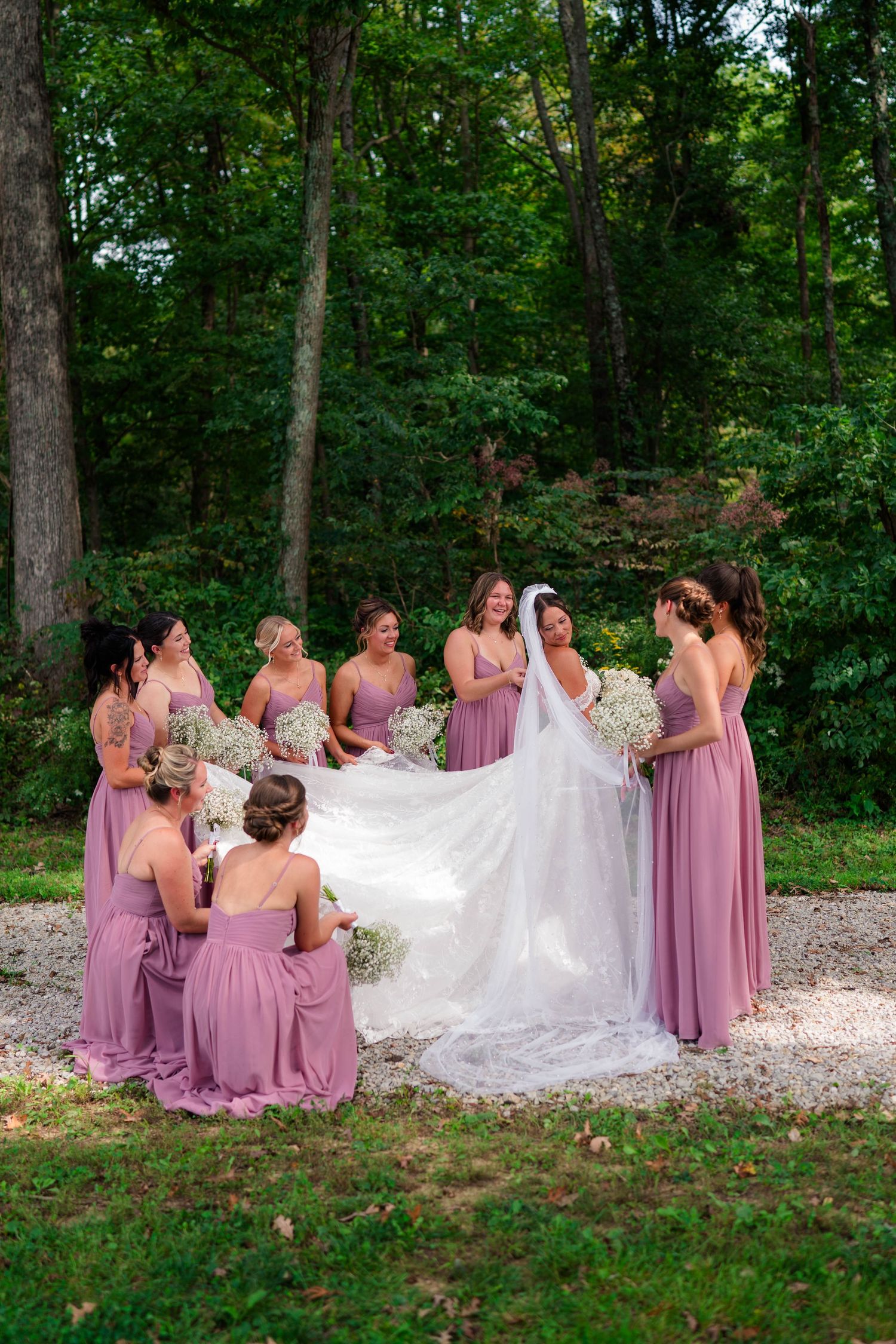 Bride surrounded by bridesmaids in dusty rose gowns holding baby's breath bouquets in wooded setting.