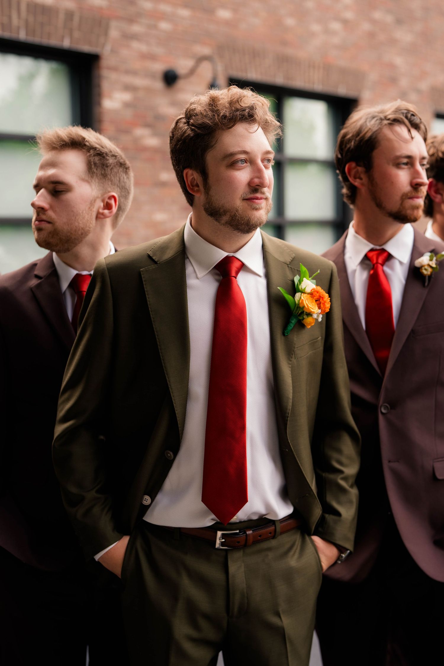 Groom in olive suit with red tie and boutonniere standing with groomsmen outside brick building.