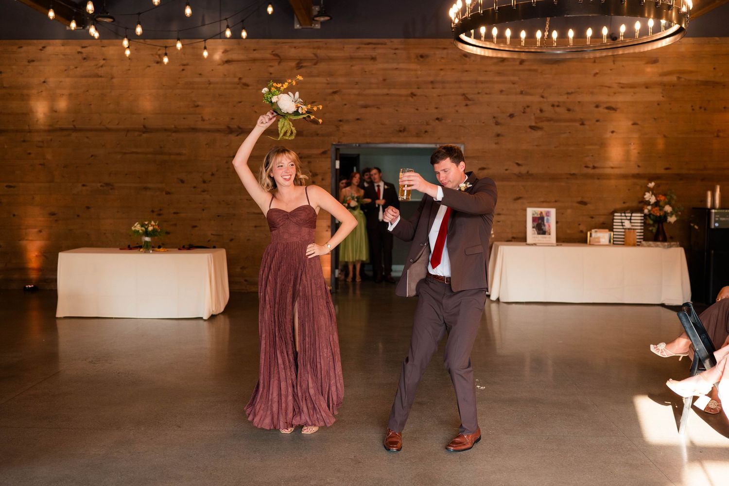 Bridesmaid and groomsman dance joyfully holding bouquet at rustic wedding reception.