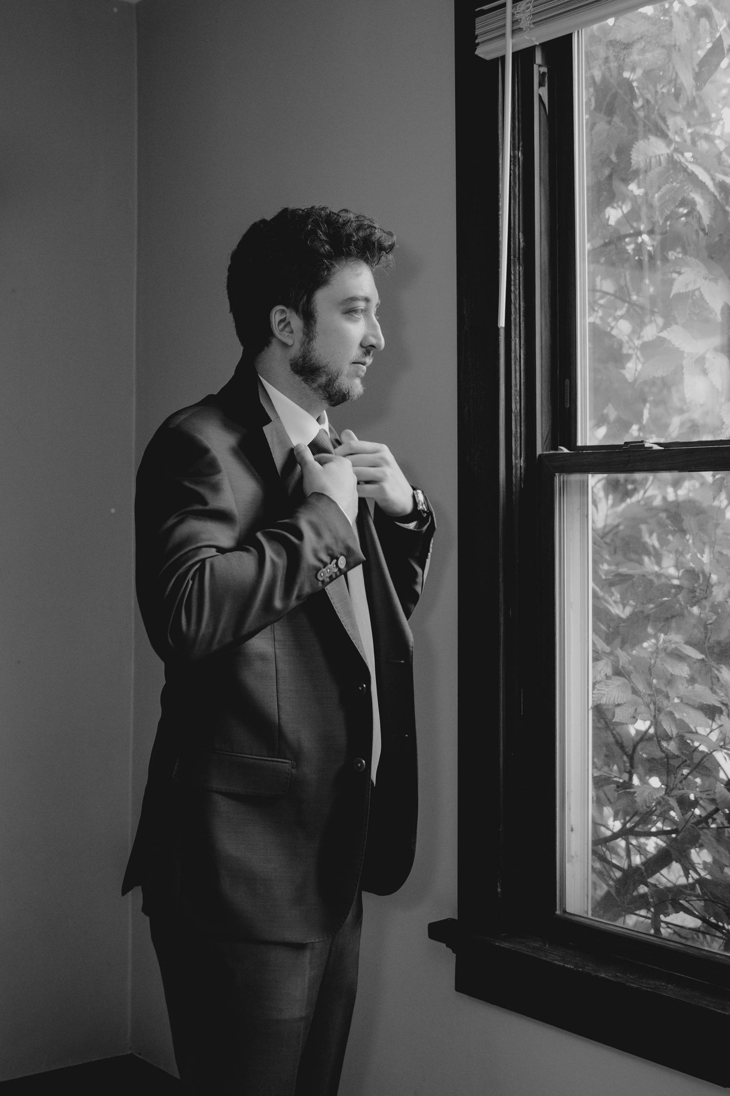 Groom in dark suit adjusting bow tie while standing by window in black and white photo.