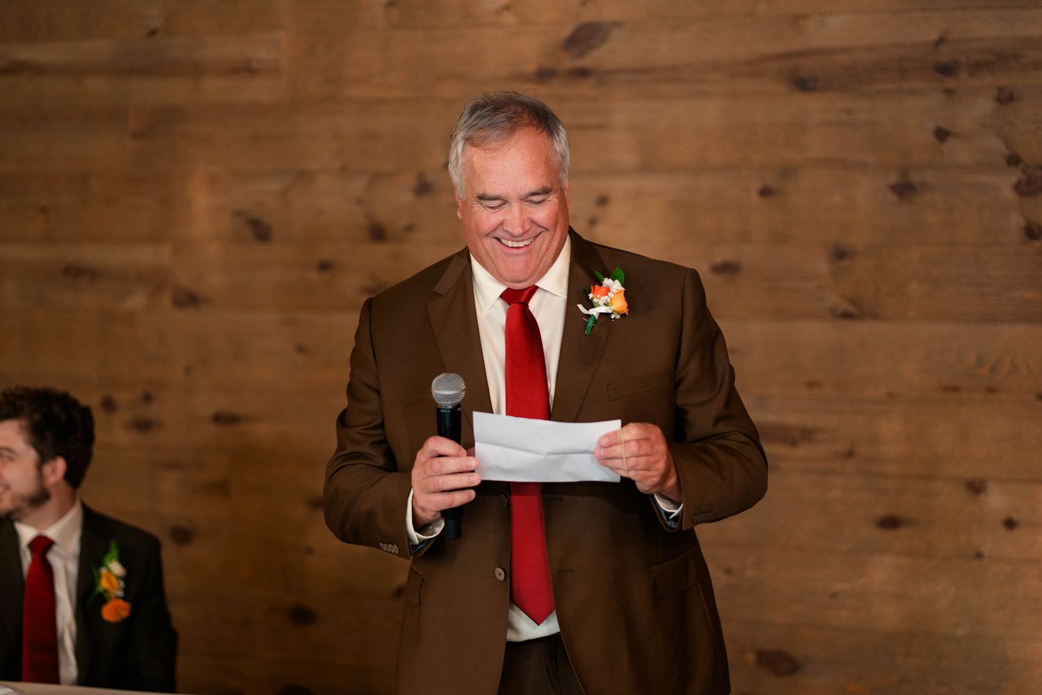 Smiling man in brown suit and red tie gives speech holding microphone at wedding.