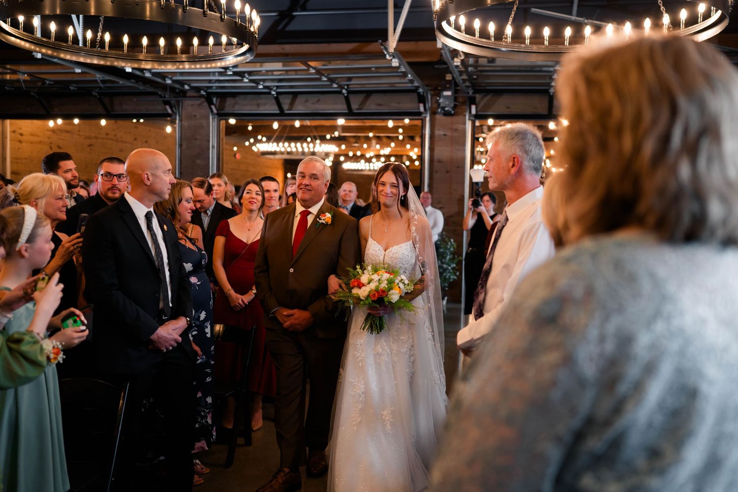 Bride walks down aisle with escorts in warmly lit venue decorated with string lights.