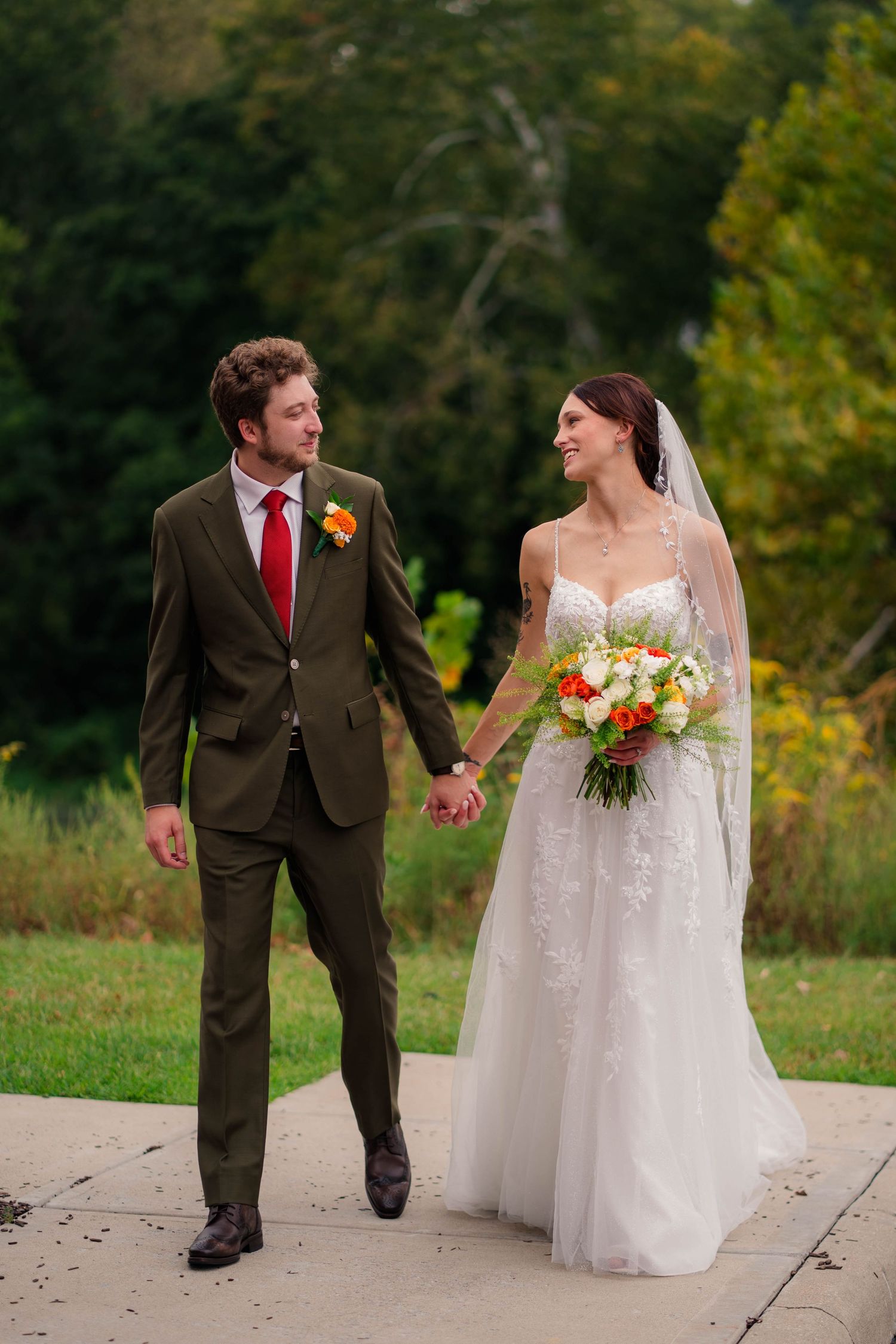 Bride and groom hold hands walking outdoors, she in lace gown with bouquet, he in olive suit with red tie.