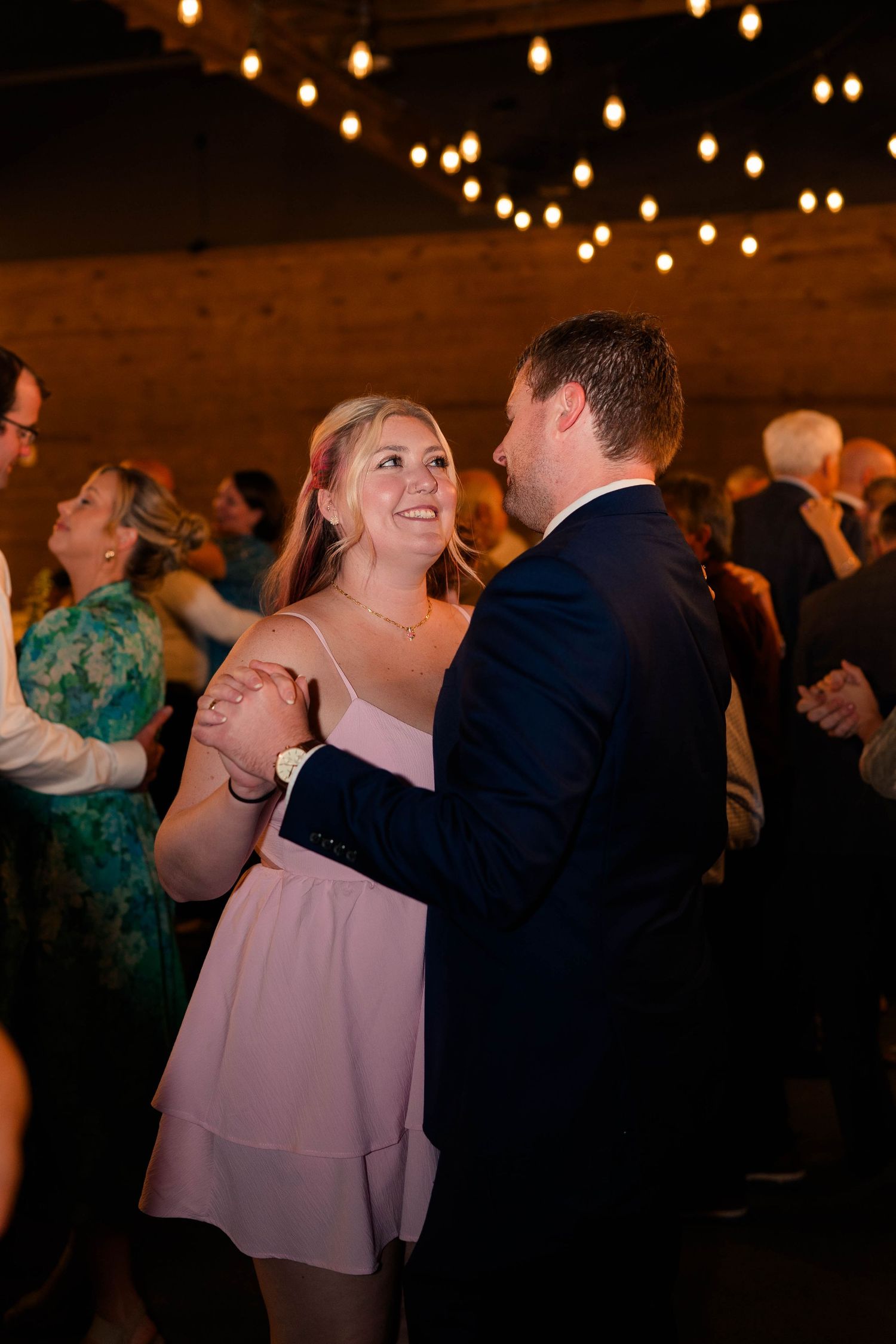 Couple dancing and smiling together under string lights at a lively wedding reception.