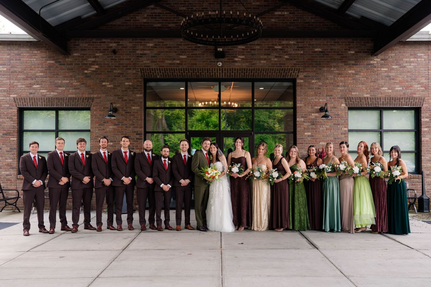 Wedding party poses outside brick venue with groomsmen in burgundy and bridesmaids in mixed green and plum gowns.