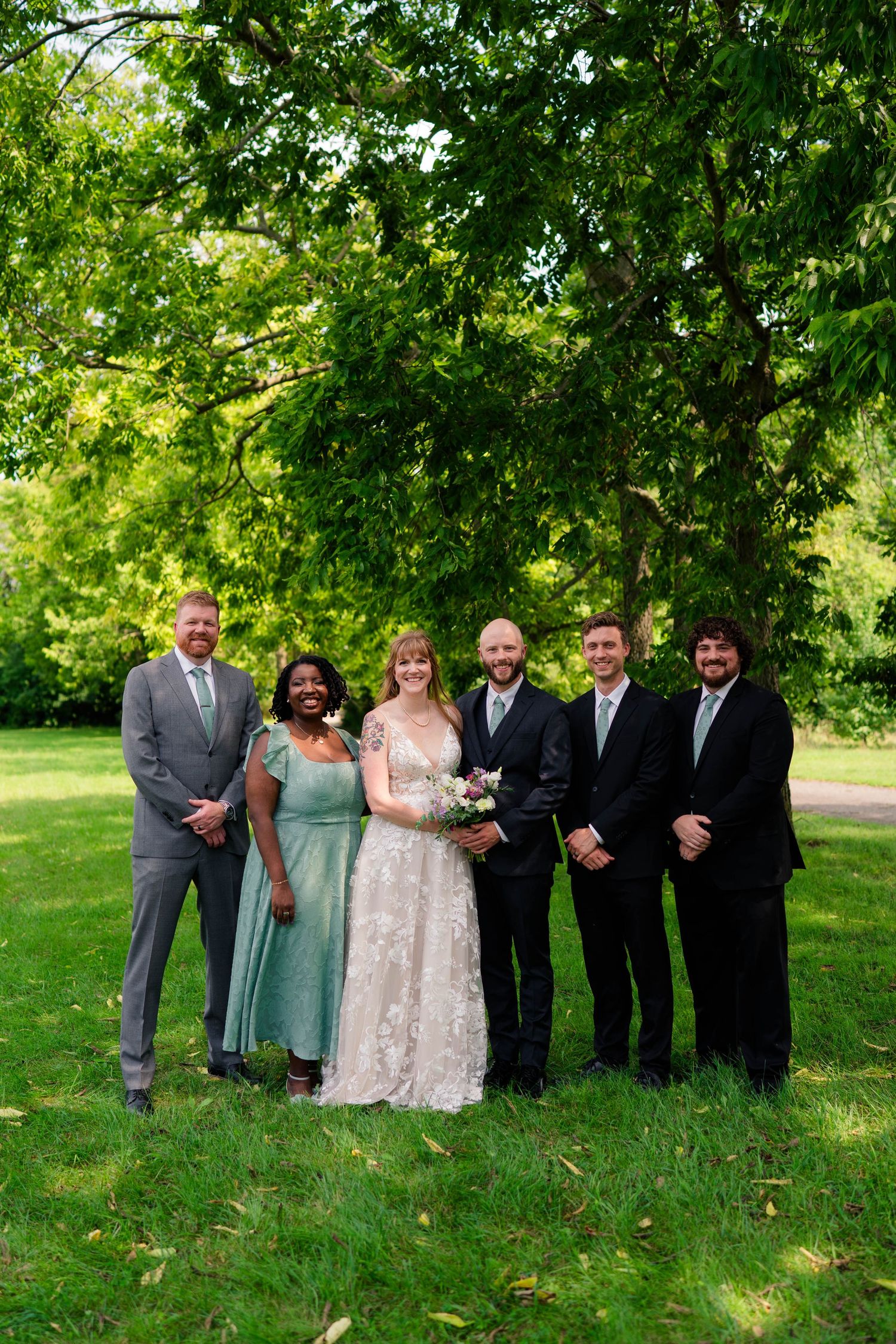 Wedding party group photo with bride, groom, and four guests smiling under lush green trees.