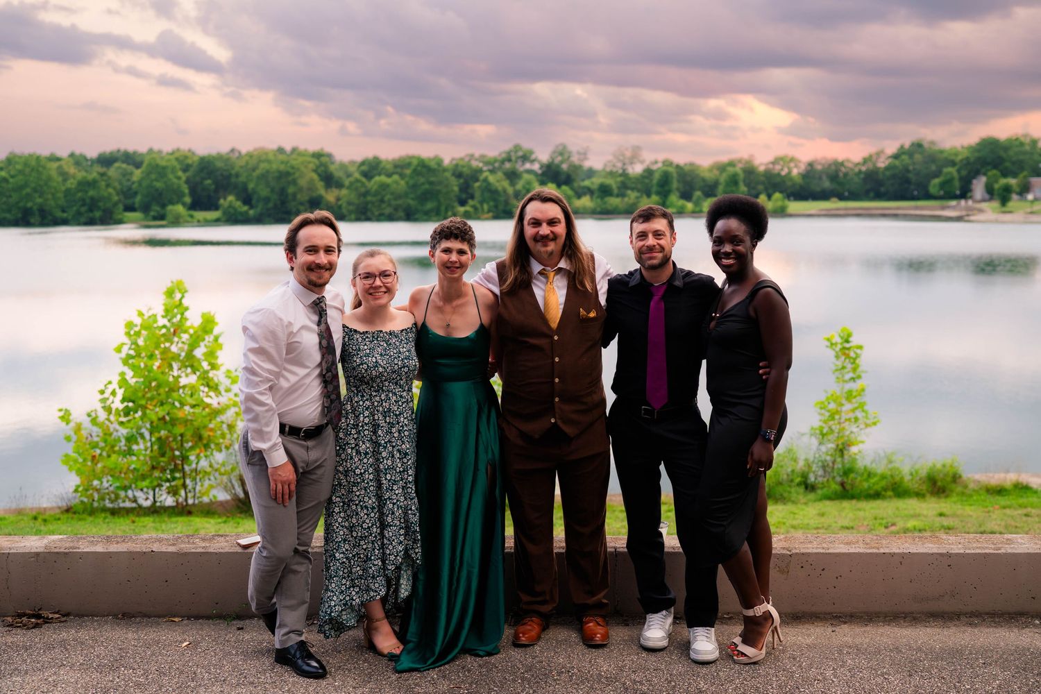 Six wedding guests posing together near a scenic lake under a colorful sunset sky.