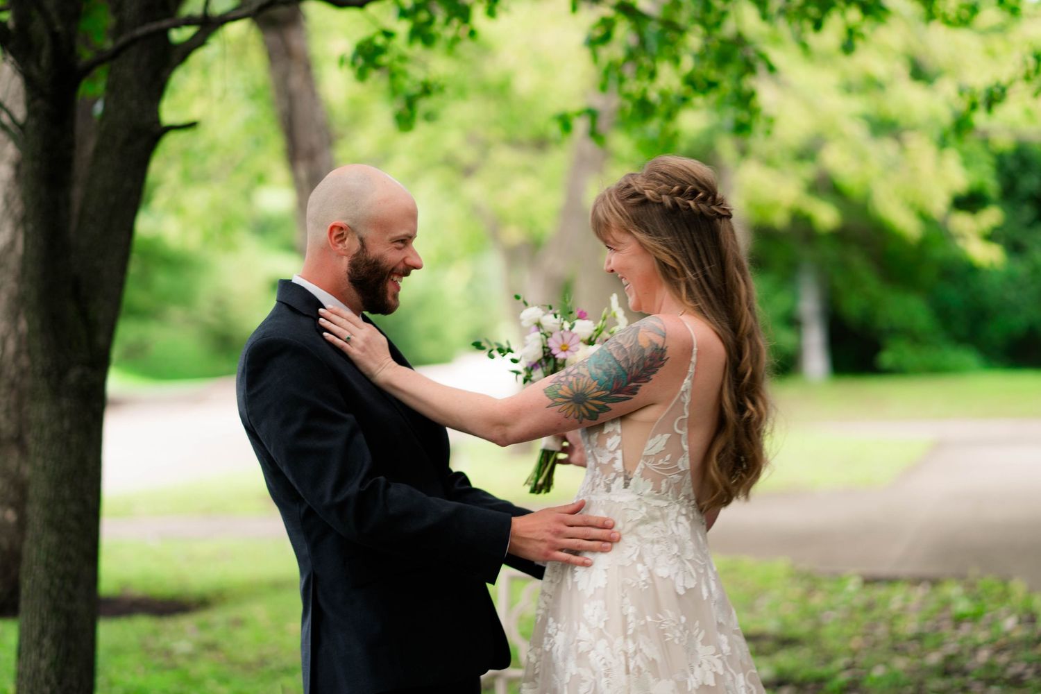 Tattooed bride and bald groom smile at each other in lush park setting.