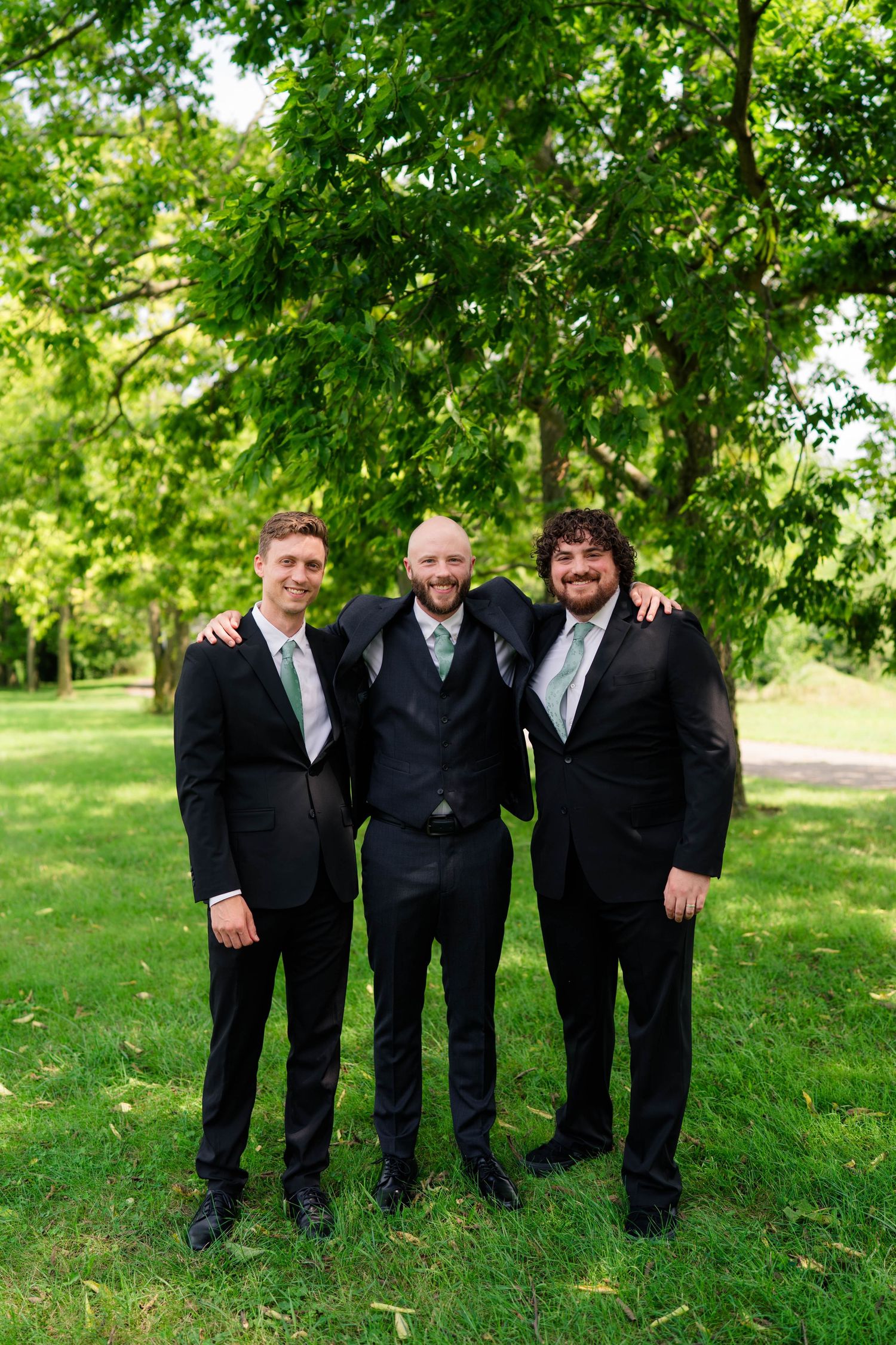 Three groomsmen in black suits with green ties posing outdoors at a wedding.
