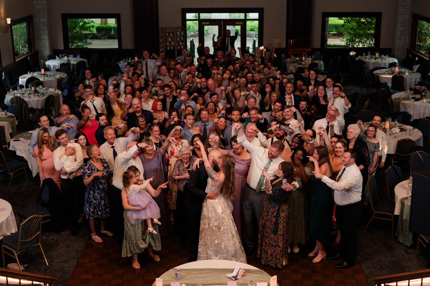Bride posing with large crowd of cheering wedding guests in a beautifully decorated venue.