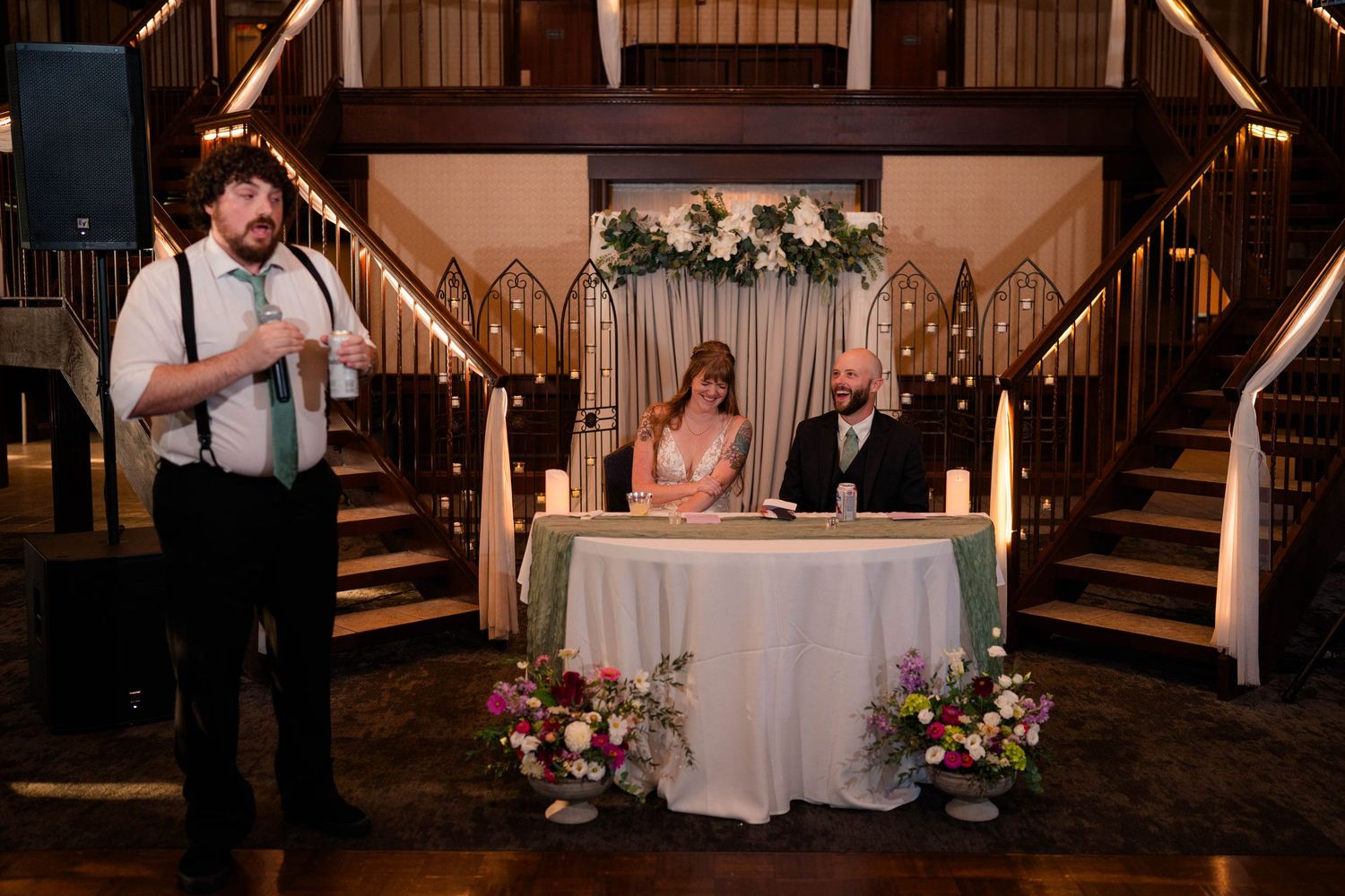 Bride and groom seated at sweetheart table with floral arch backdrop while officiant delivers speech at wedding reception.