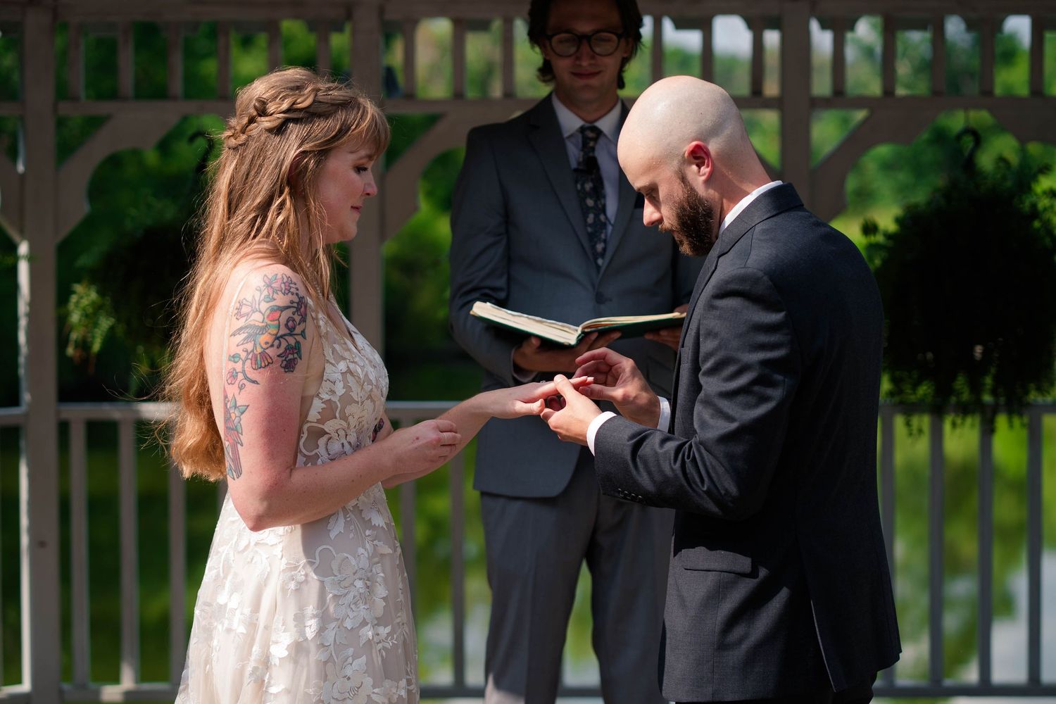Groom places wedding ring on brides finger during outdoor ceremony beneath a white gazebo.