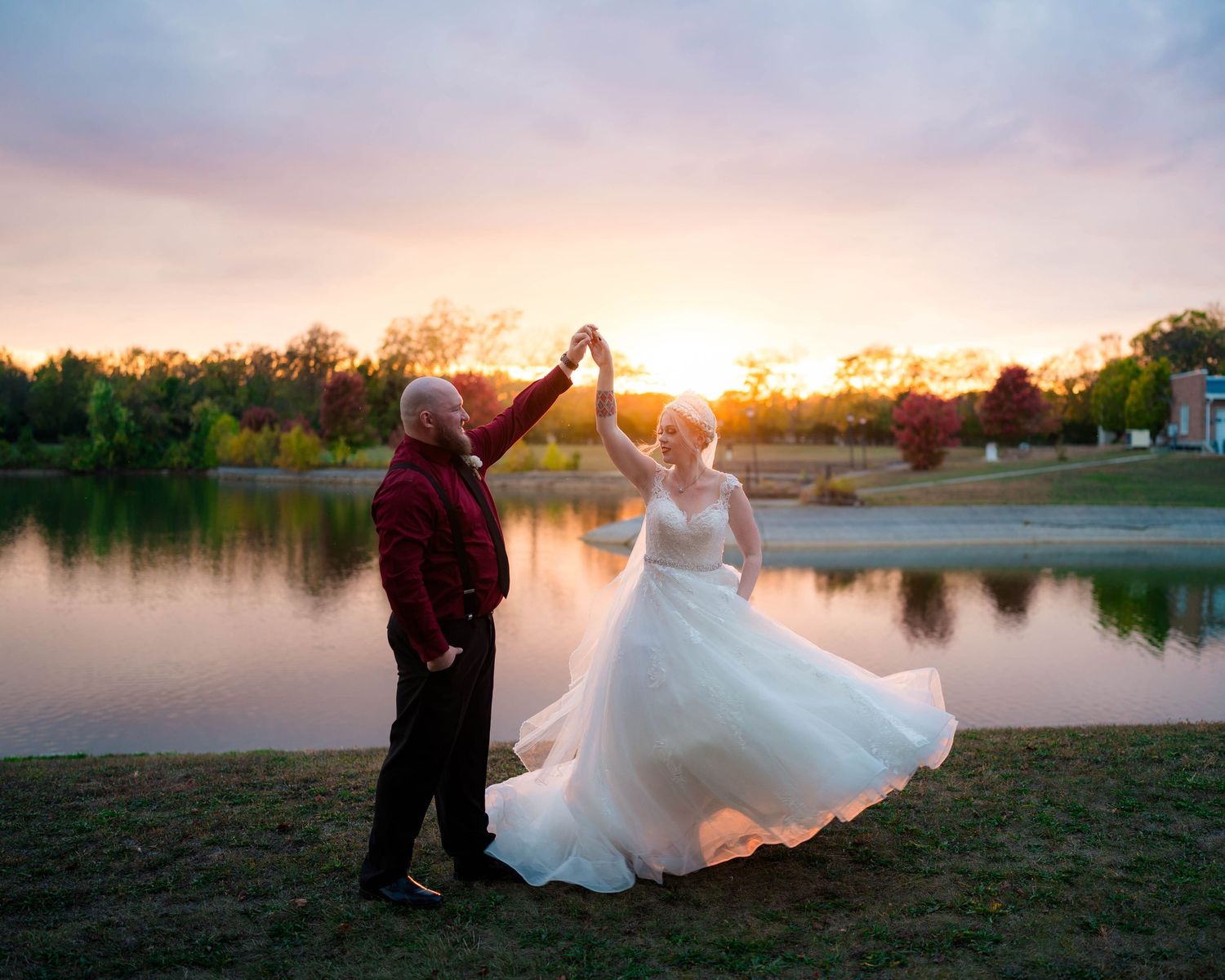 Groom twirls bride in a white ball gown at sunset beside a serene reflective lake.