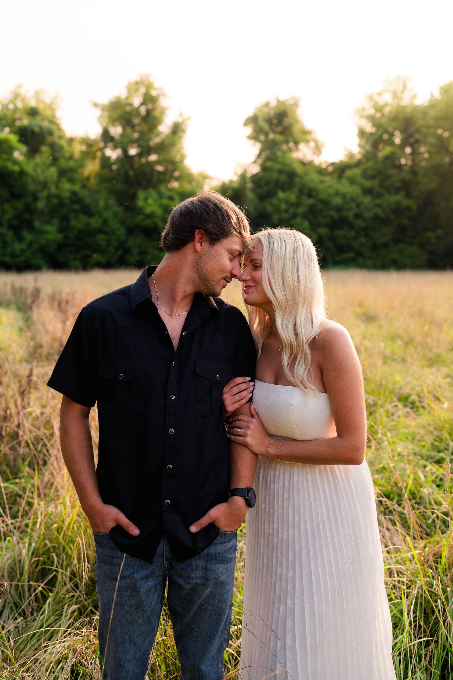 A couple shares a romantic forehead touch in a golden field at sunset, man in black shirt and woman in white dress.