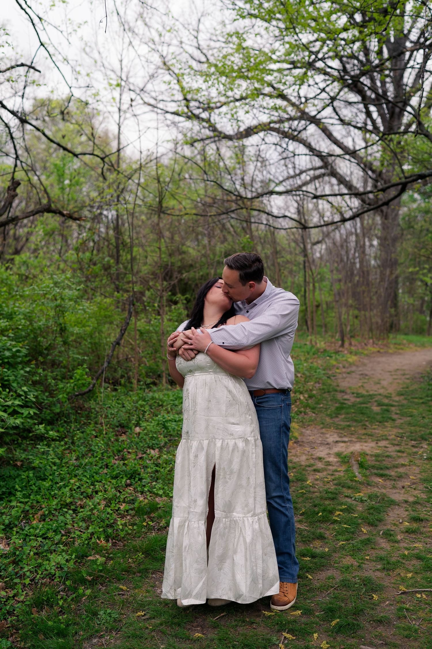 Couple sharing a tender kiss embrace on a wooded nature trail surrounded by spring foliage.