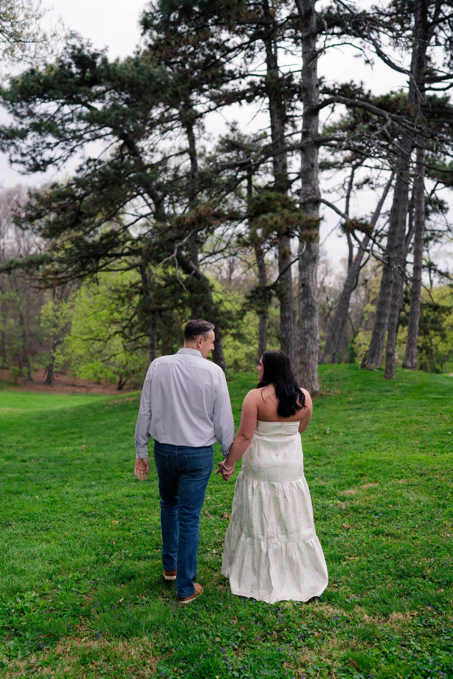 Couple holding hands walking through lush green park toward tall pine trees, woman in white dress and man in jeans.