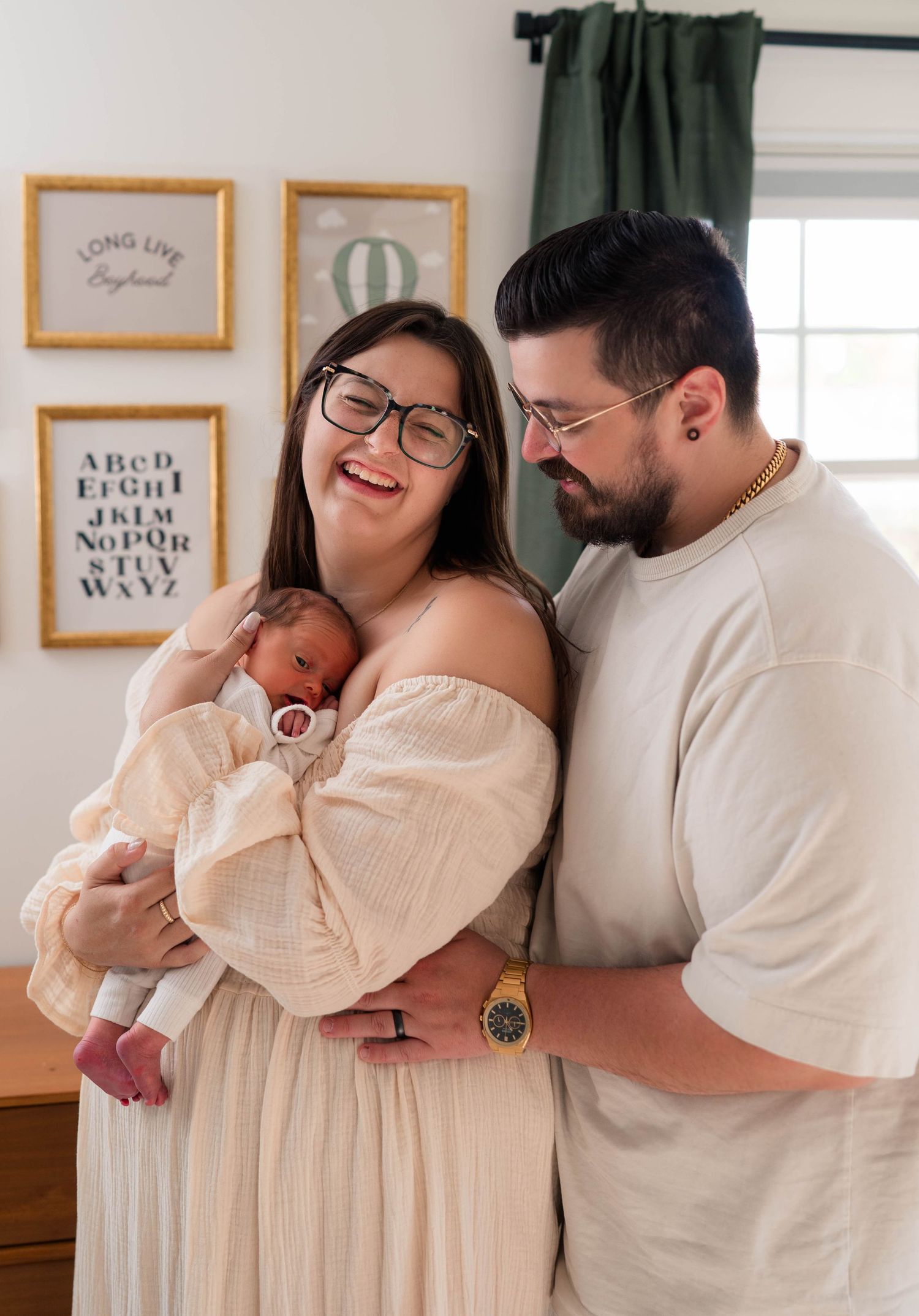 Happy couple wearing glasses laughs together while cradling their swaddled newborn baby at home.