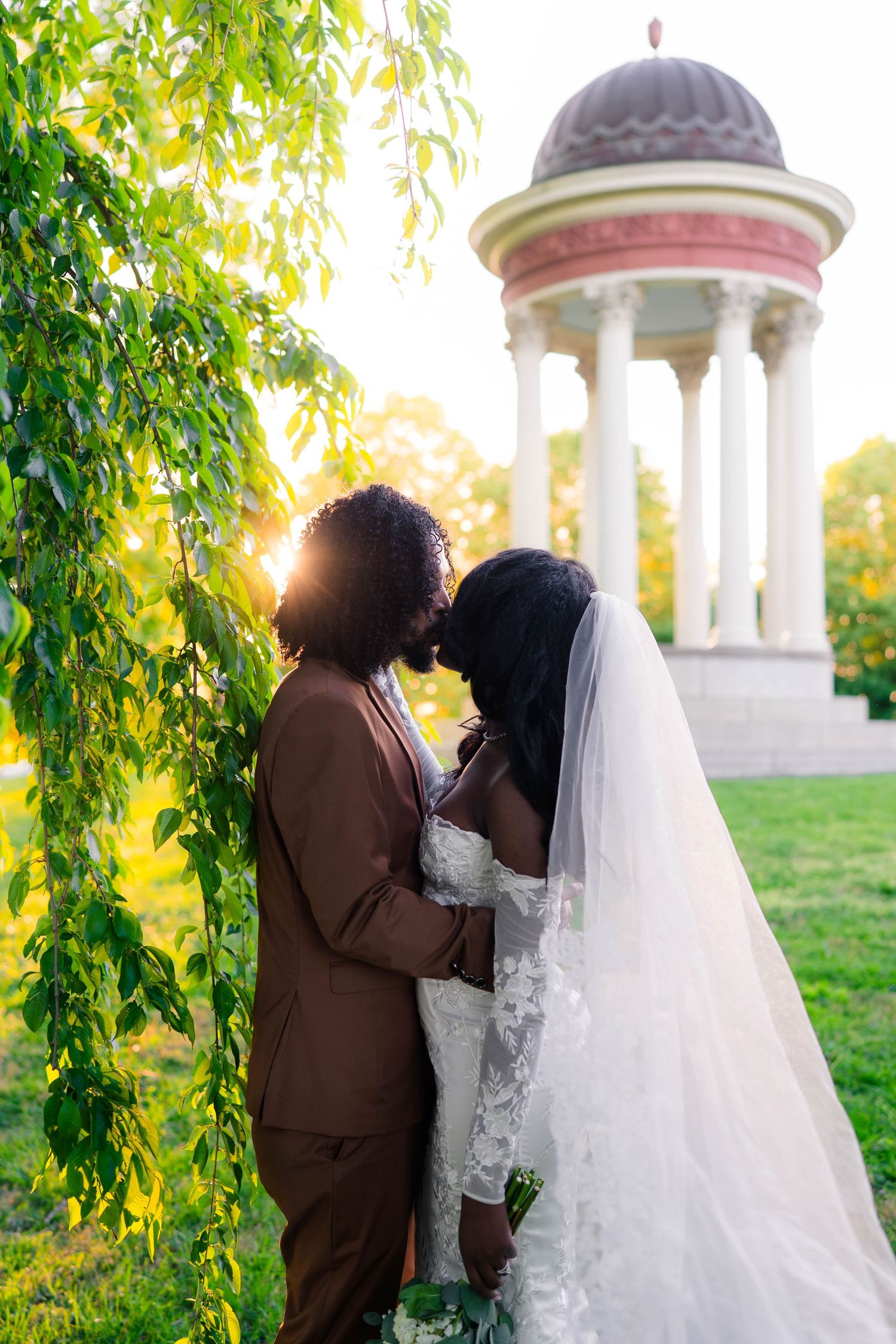 Black couple shares romantic kiss in wedding attire near weeping willow with sunlit gazebo backdrop.