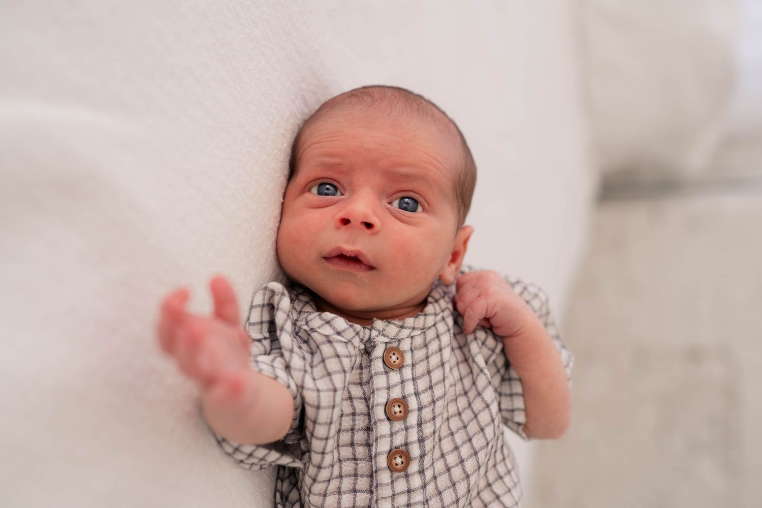 Newborn baby with blue eyes in checkered onesie lying on white blanket, reaching out tiny hand.