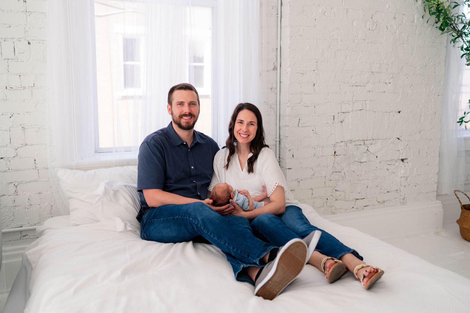 Smiling couple sitting on a white bed holding their newborn baby indoors.