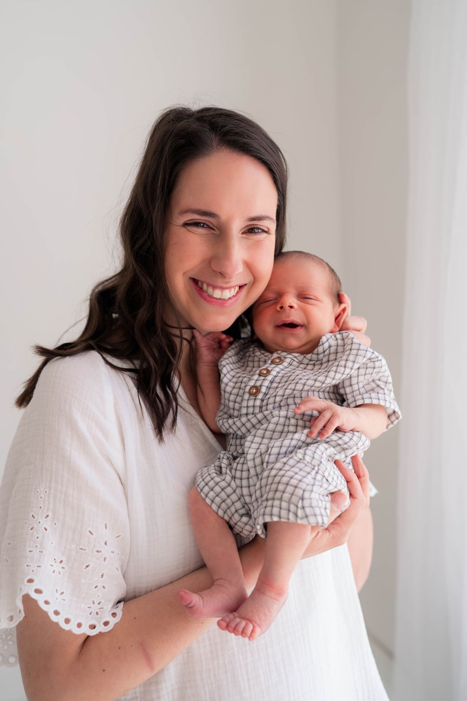 Smiling mother in white dress holds her newborn baby in a checkered onesie against a bright white background.