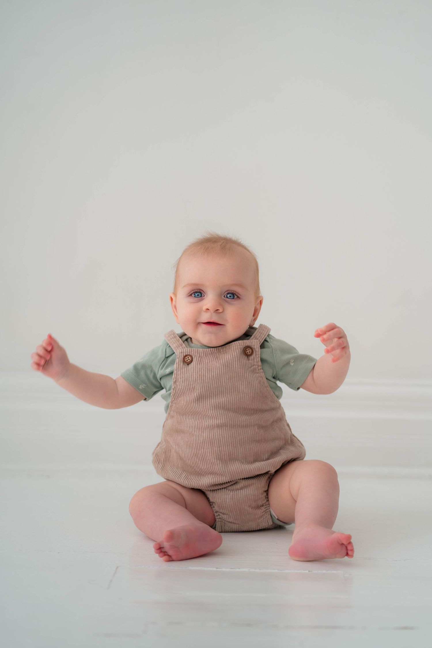 Smiling baby with blue eyes sitting on white floor wearing green shirt and brown linen overalls.