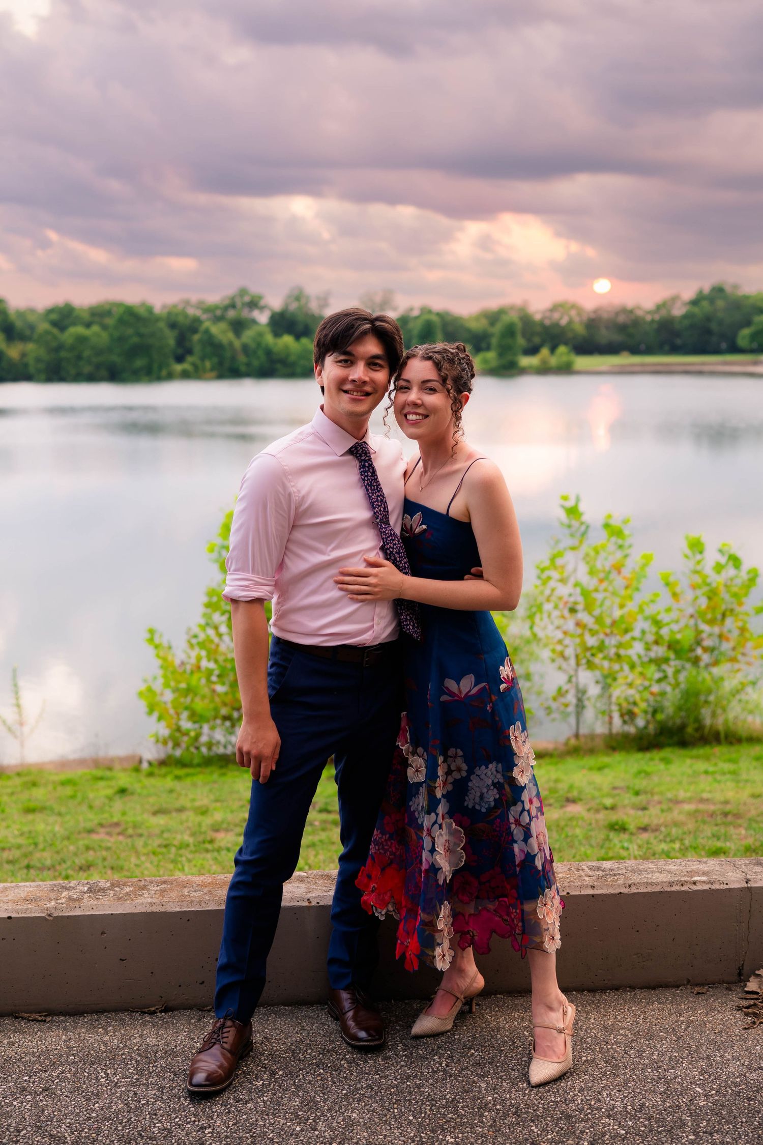 Young couple in formal attire embraces and smiles by a lake at a colorful sunset.
