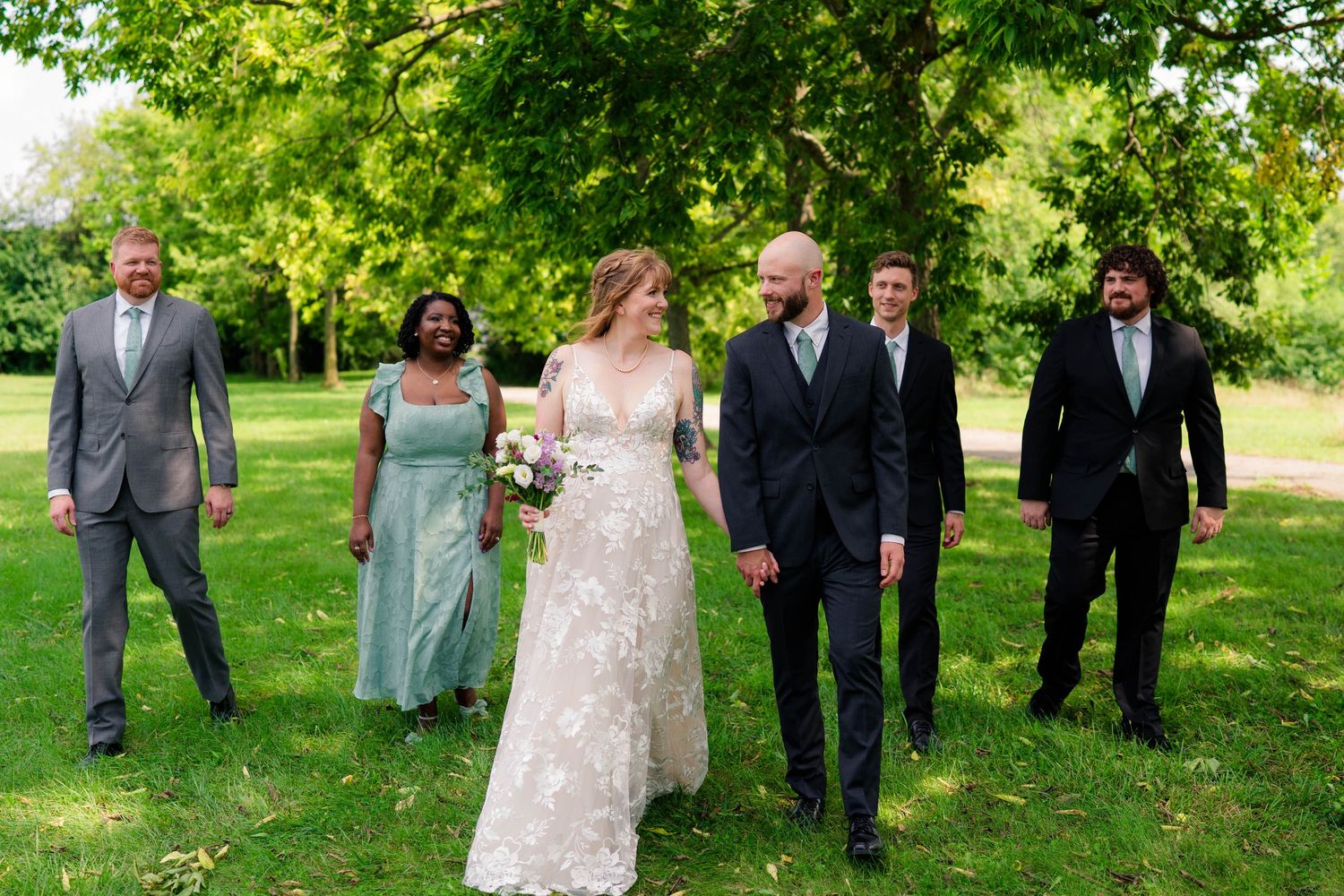 Bride and groom walk with wedding party through green park, bride in floral lace gown, attendants in navy and sage.