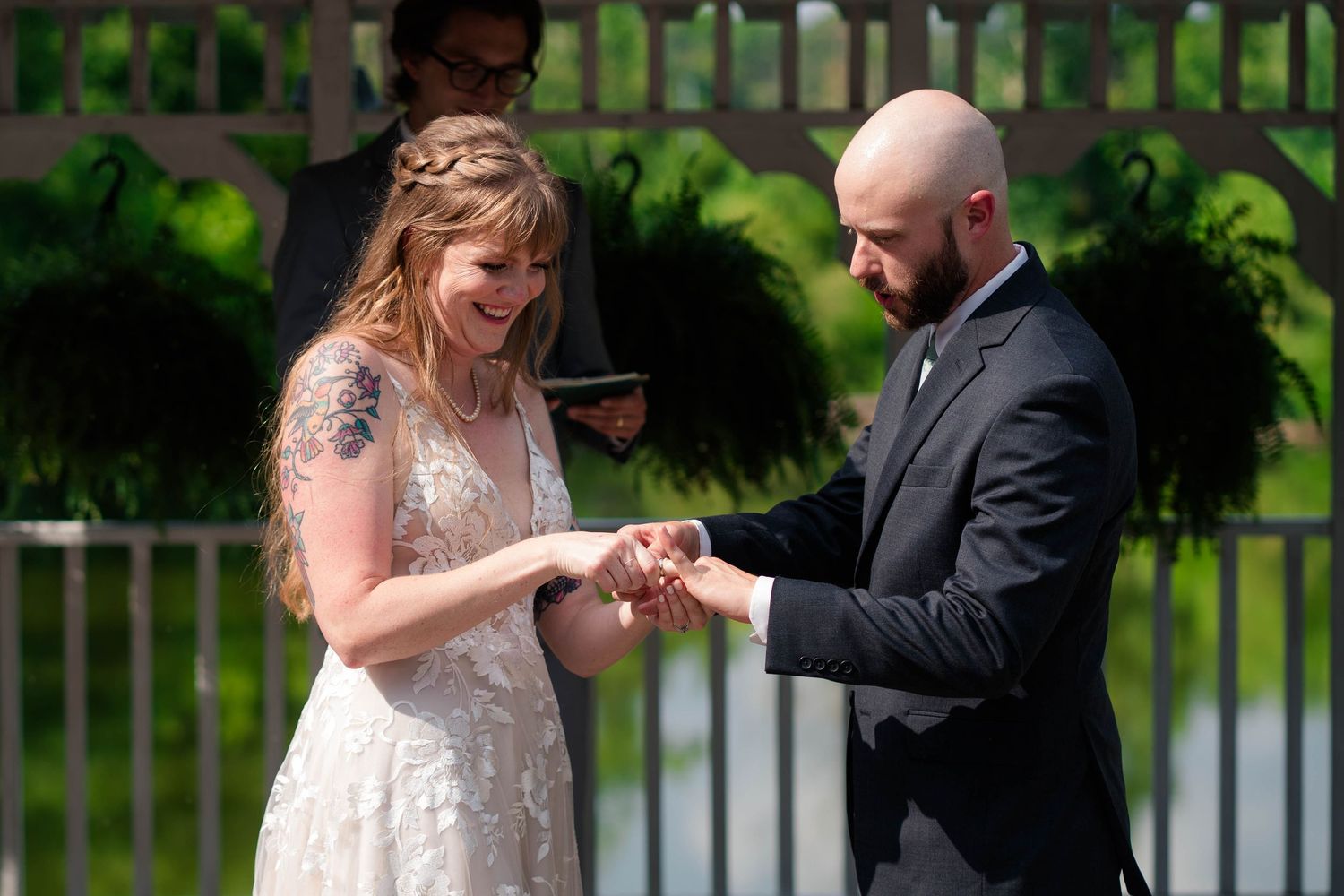 Laughing bride with tattoos receives wedding ring from bald groom during joyful outdoor ceremony exchange.