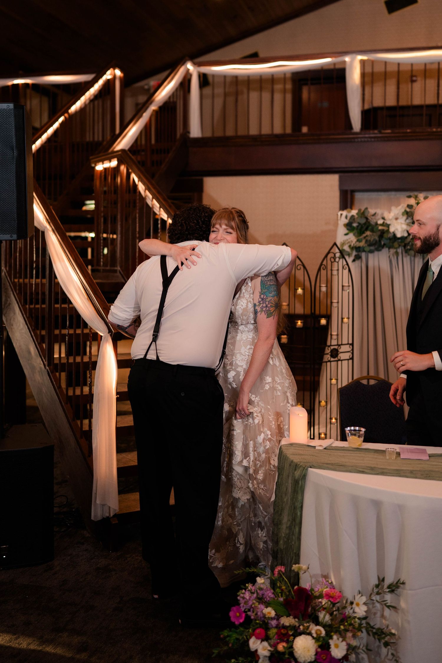 Bride hugs emotional guest near sweetheart table at warmly lit indoor wedding reception.