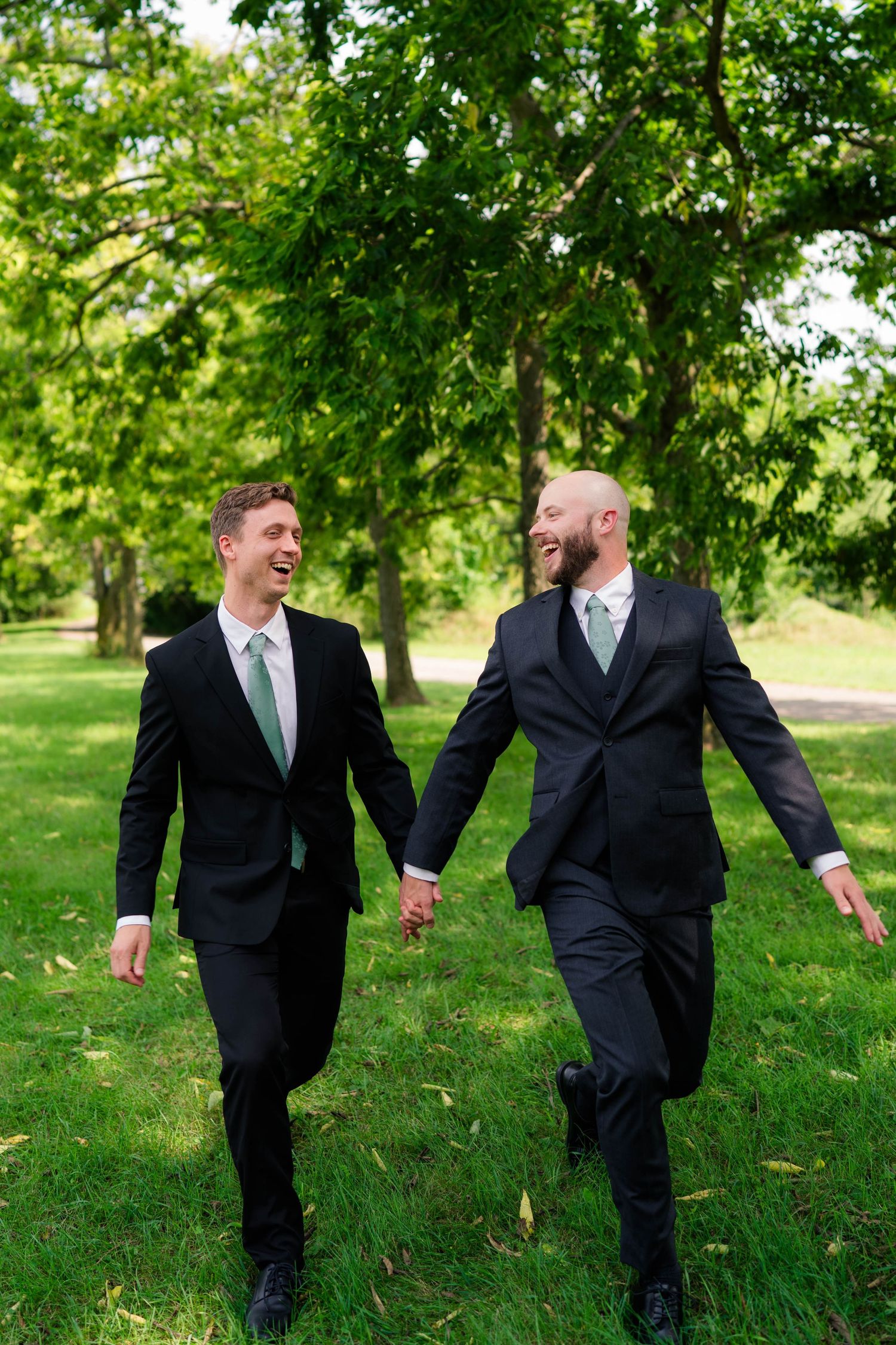 Two laughing groomsmen in black suits hold hands while walking across green grass in sunny park.