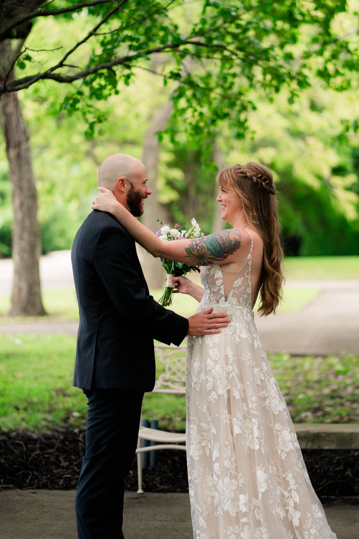 Bride and groom share a joyful first look moment outdoors, bride in lace gown holding bouquet.