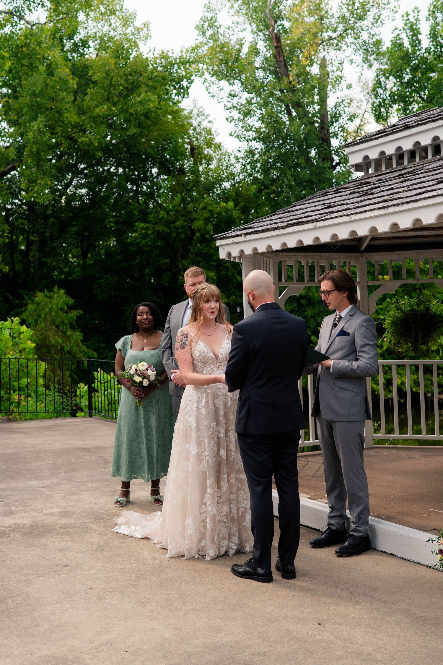 Bride smiles during outdoor wedding ceremony near white gazebo with bridesmaids and groomsmen standing nearby.