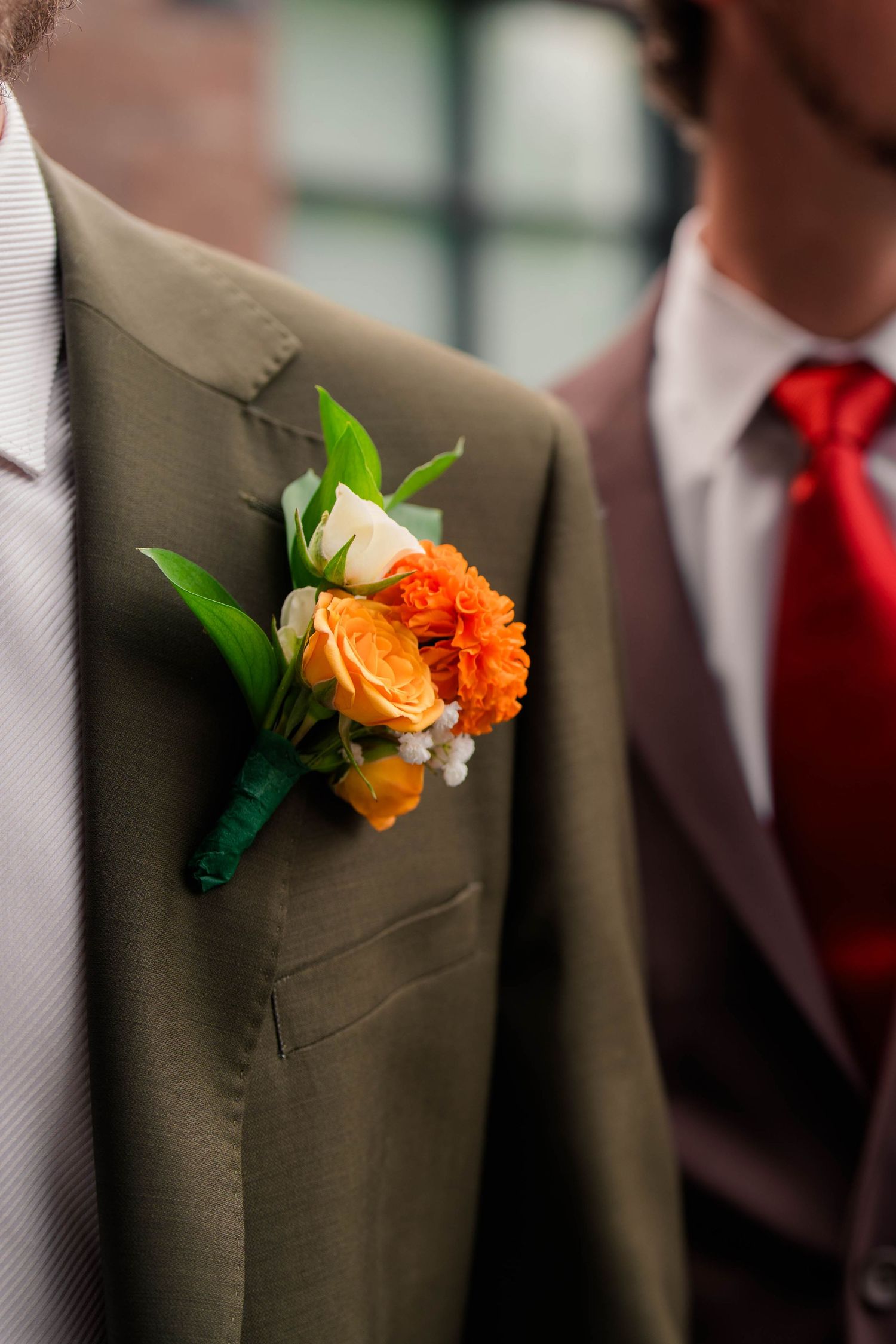 Close-up of orange rose boutonniere on olive green groom suit at wedding ceremony.