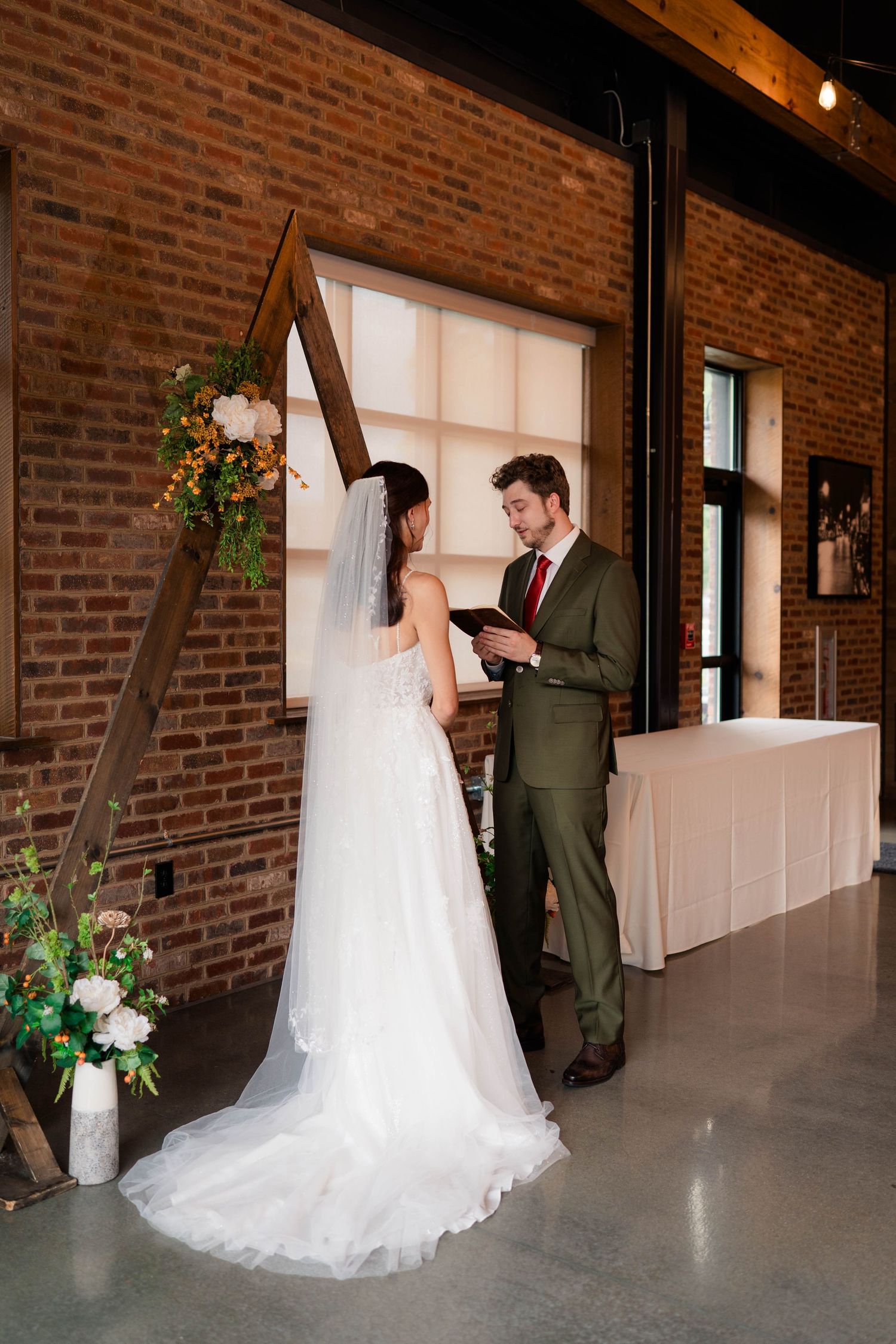 Bride and groom exchange vows beneath a wooden triangular arch adorned with flowers in a rustic brick venue.