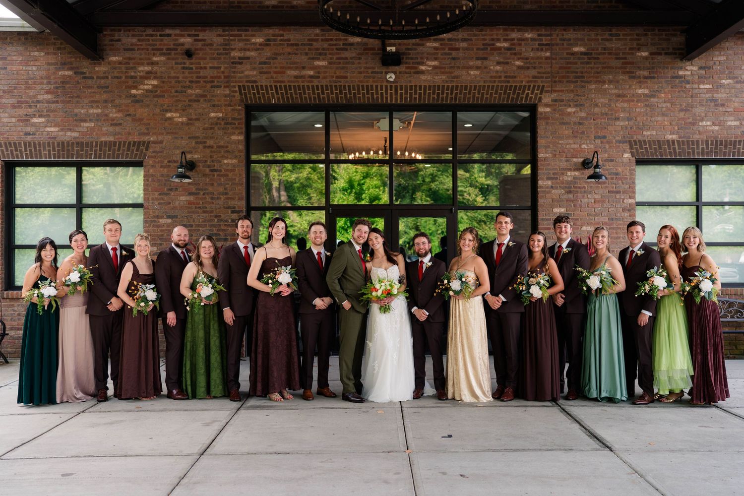 Large wedding party poses outside brick venue, bridesmaids in green and burgundy gowns, groomsmen in brown suits.