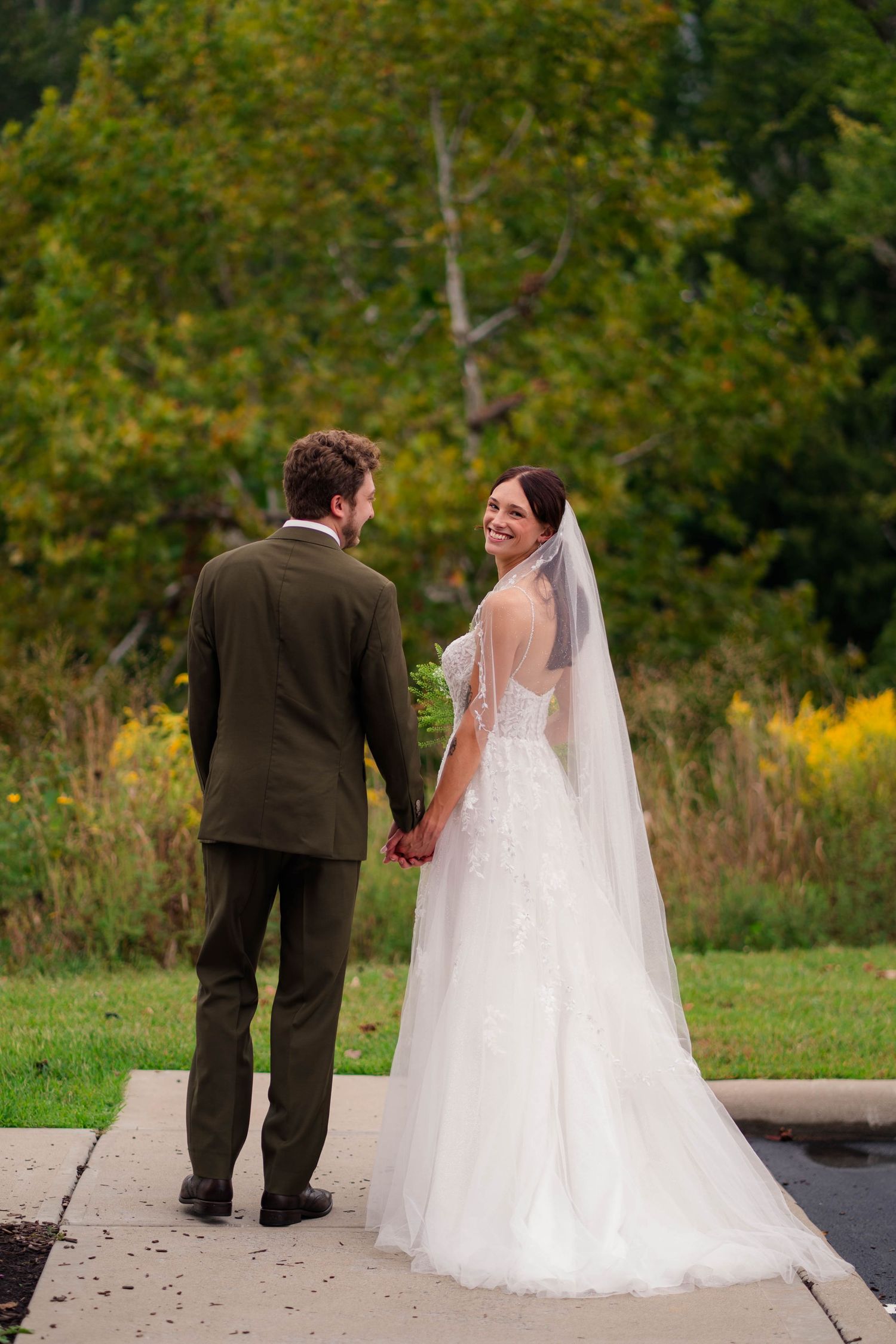 Smiling bride in lace gown and veil looks back while holding hands with groom outdoors in autumn setting.