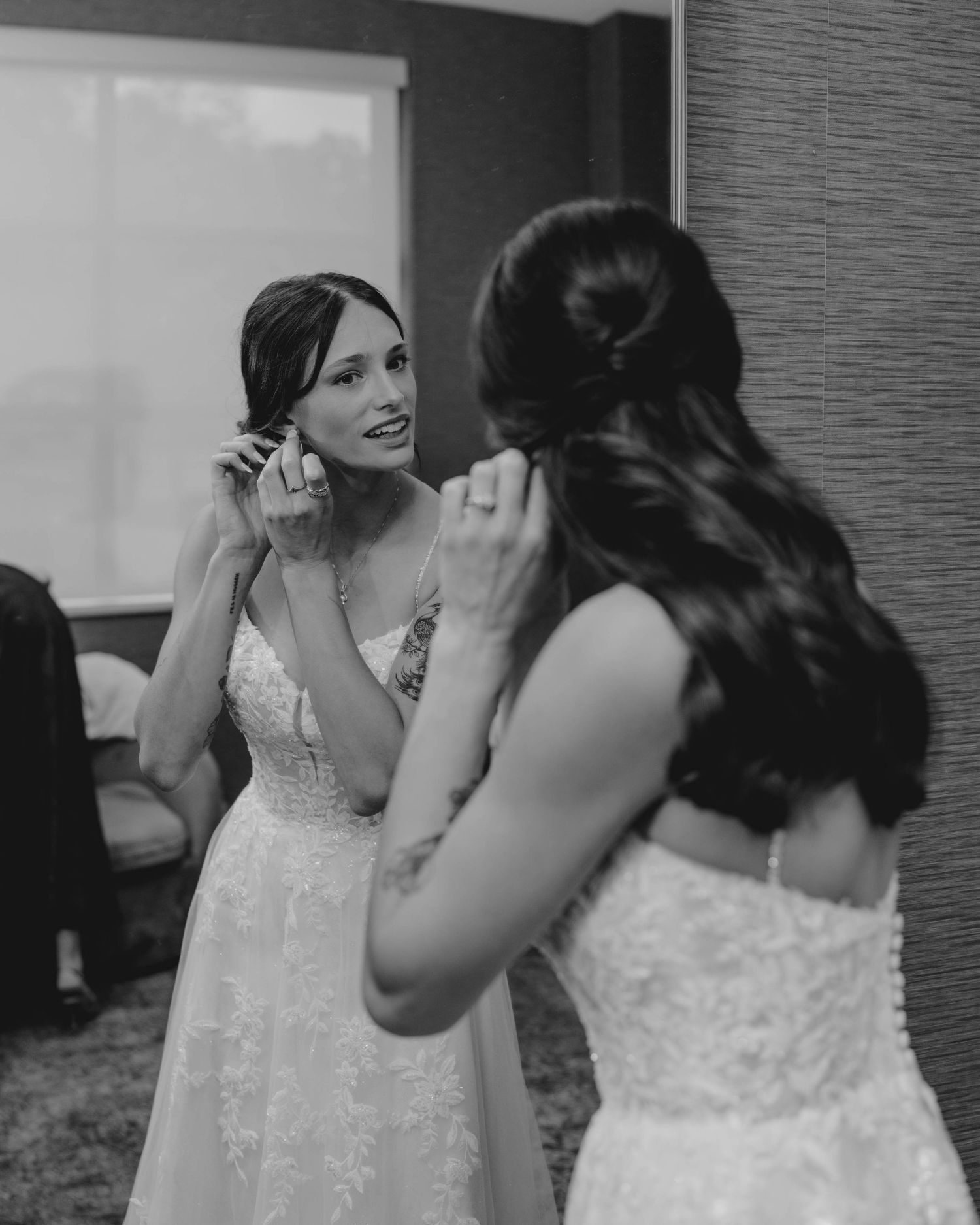 Bride putting on earrings while looking in mirror on wedding day in black and white photo.