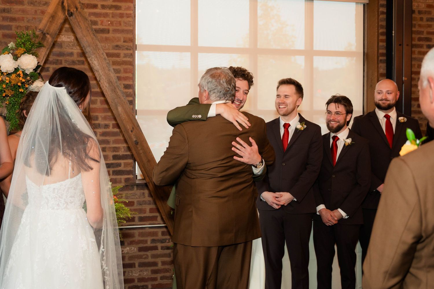 Groom embraces guest during wedding ceremony at rustic brick venue with groomsmen smiling nearby.