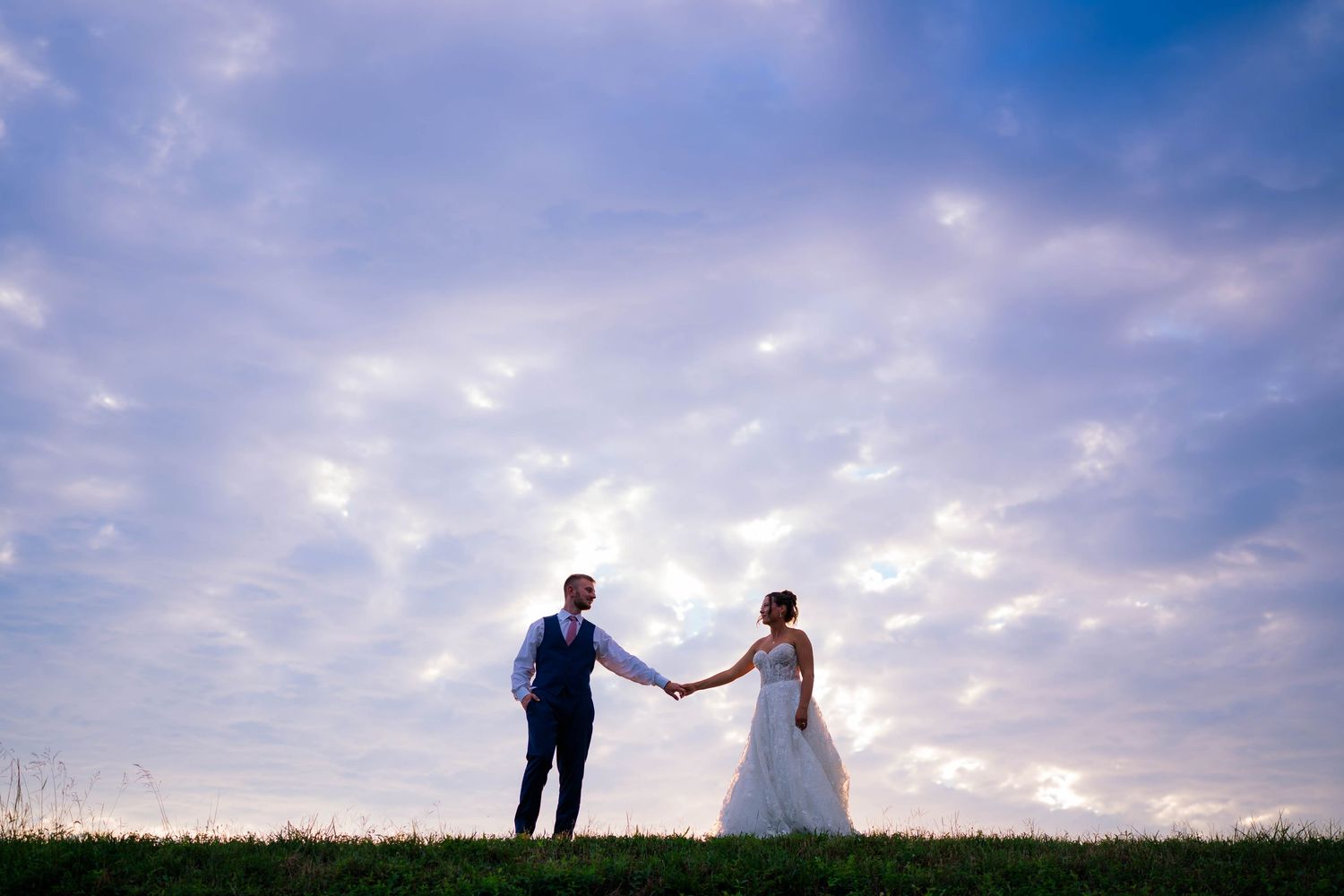 Bride and groom hold hands silhouetted against a dramatic purple sunset sky.